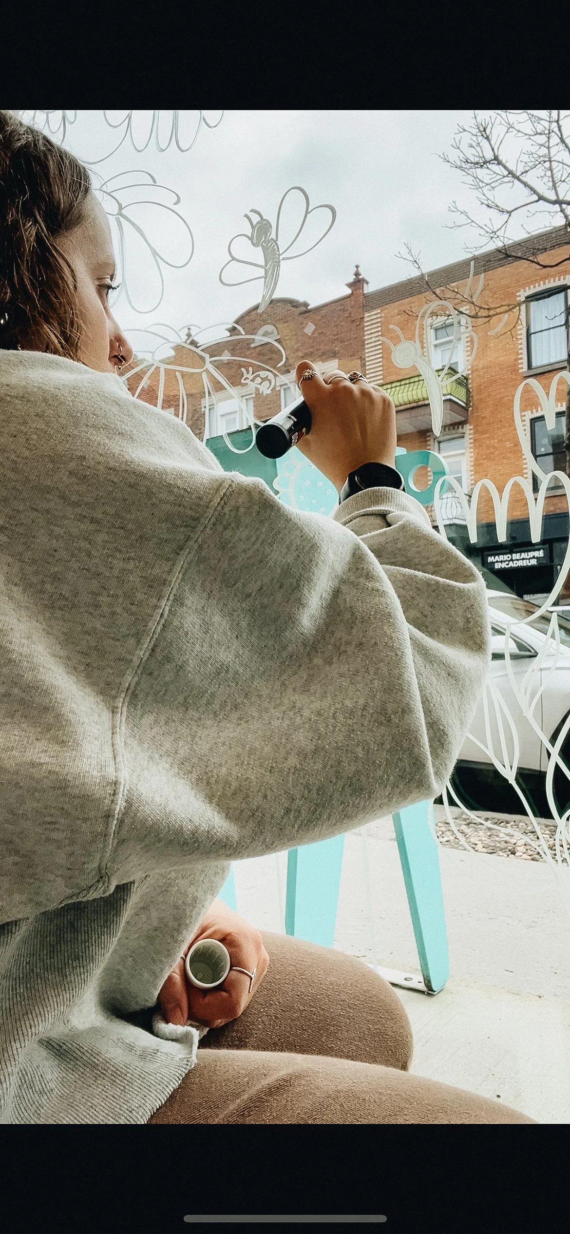 Une femme regarde par une grande vitre décorée de dessins de fleurs et d'insectes, en utilisant un marqueur noir pour ajouter à la décoration.