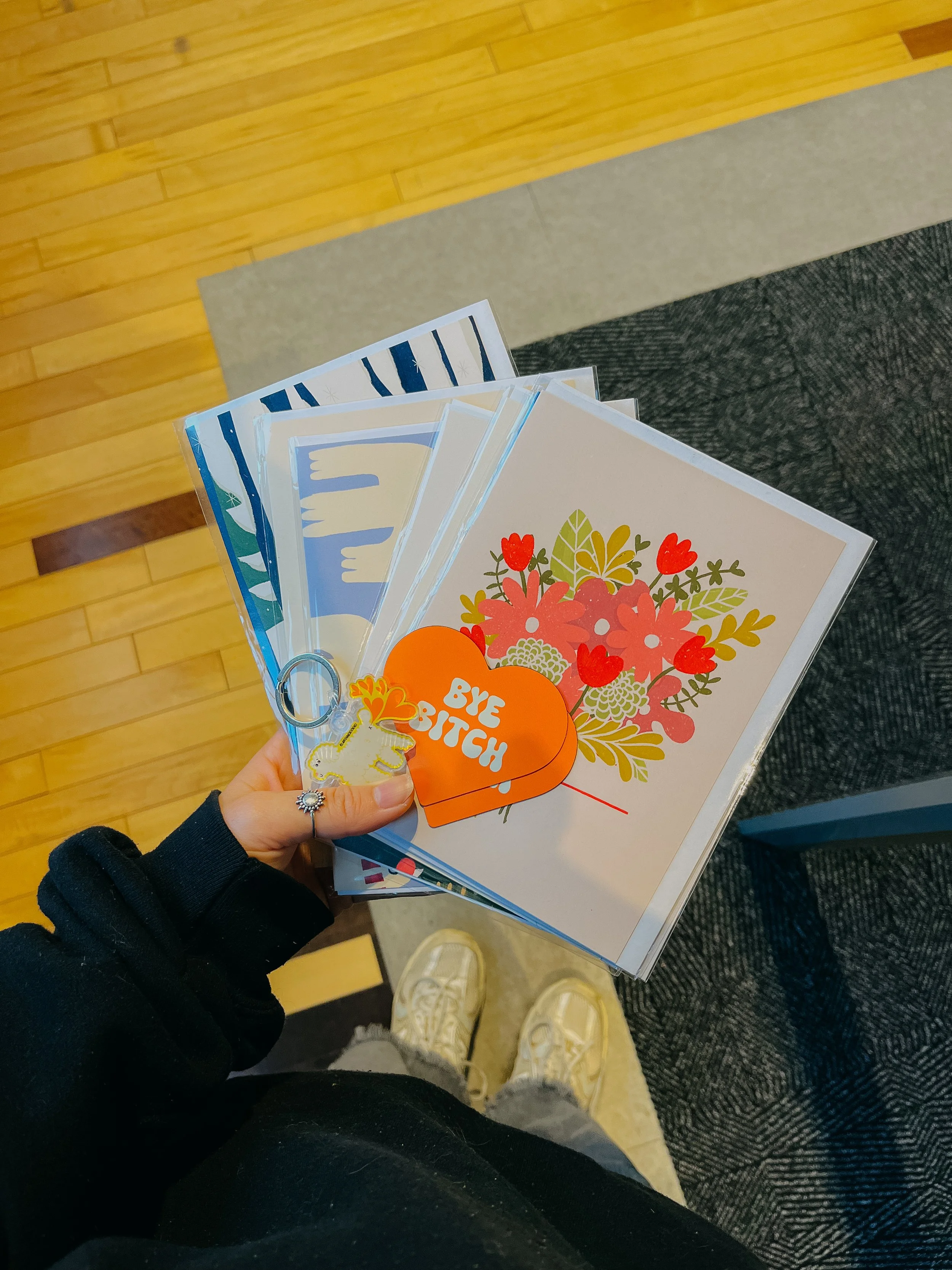 Main tenant un lot de dessins ou de cartes avec divers motifs floraux et la phrase 'Bye Bitch', vue du dessus, avec des chaussures blanches et un jean gris au sol, et un sol en parquet et tapis en arrière-plan.