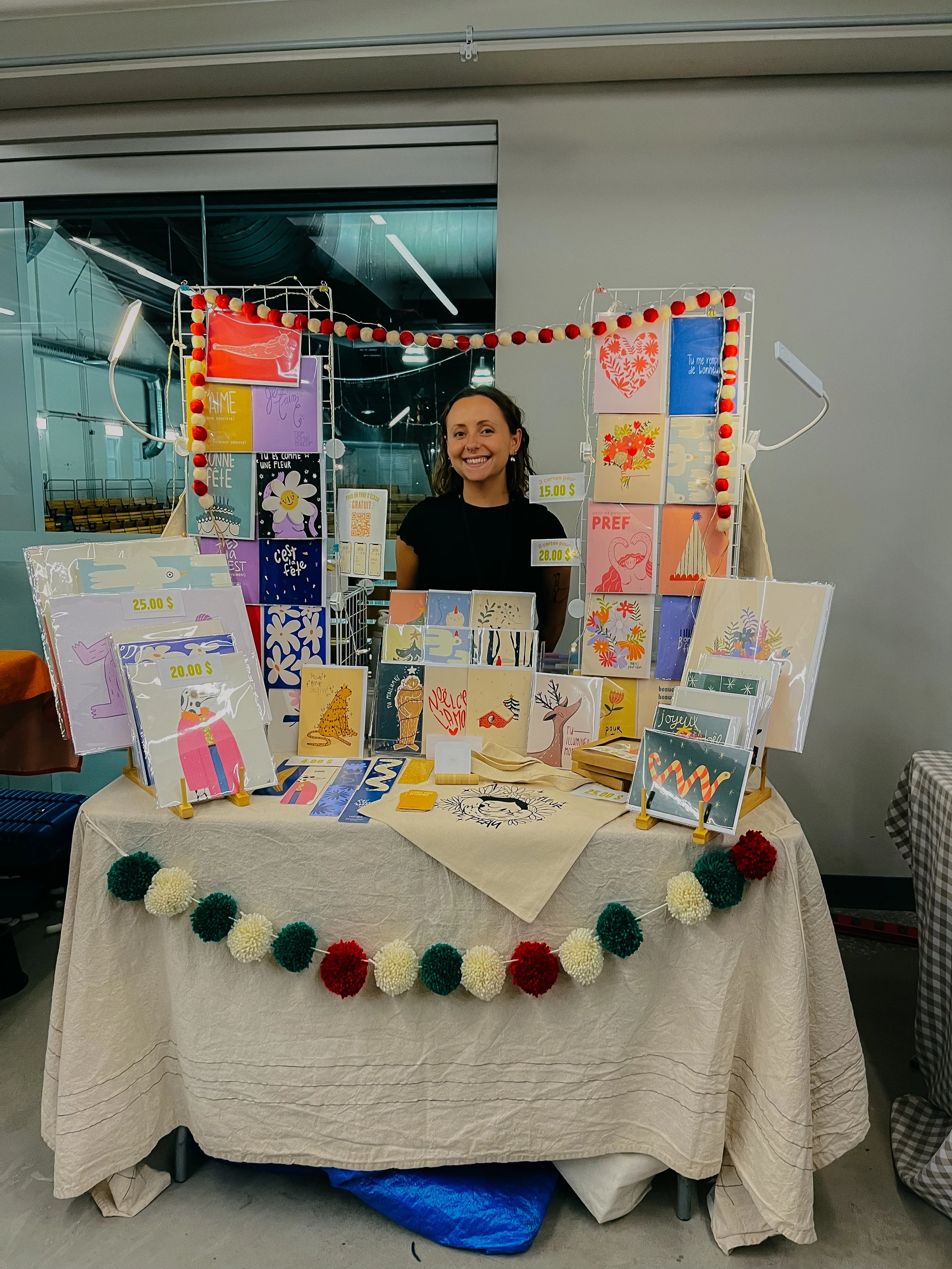Un stand avec des cartes de vœux colorées et décorations, un femme souriante derrière la table, décoré avec une guirlande de pompons dans les couleurs tricolores, dans un espace intérieur.