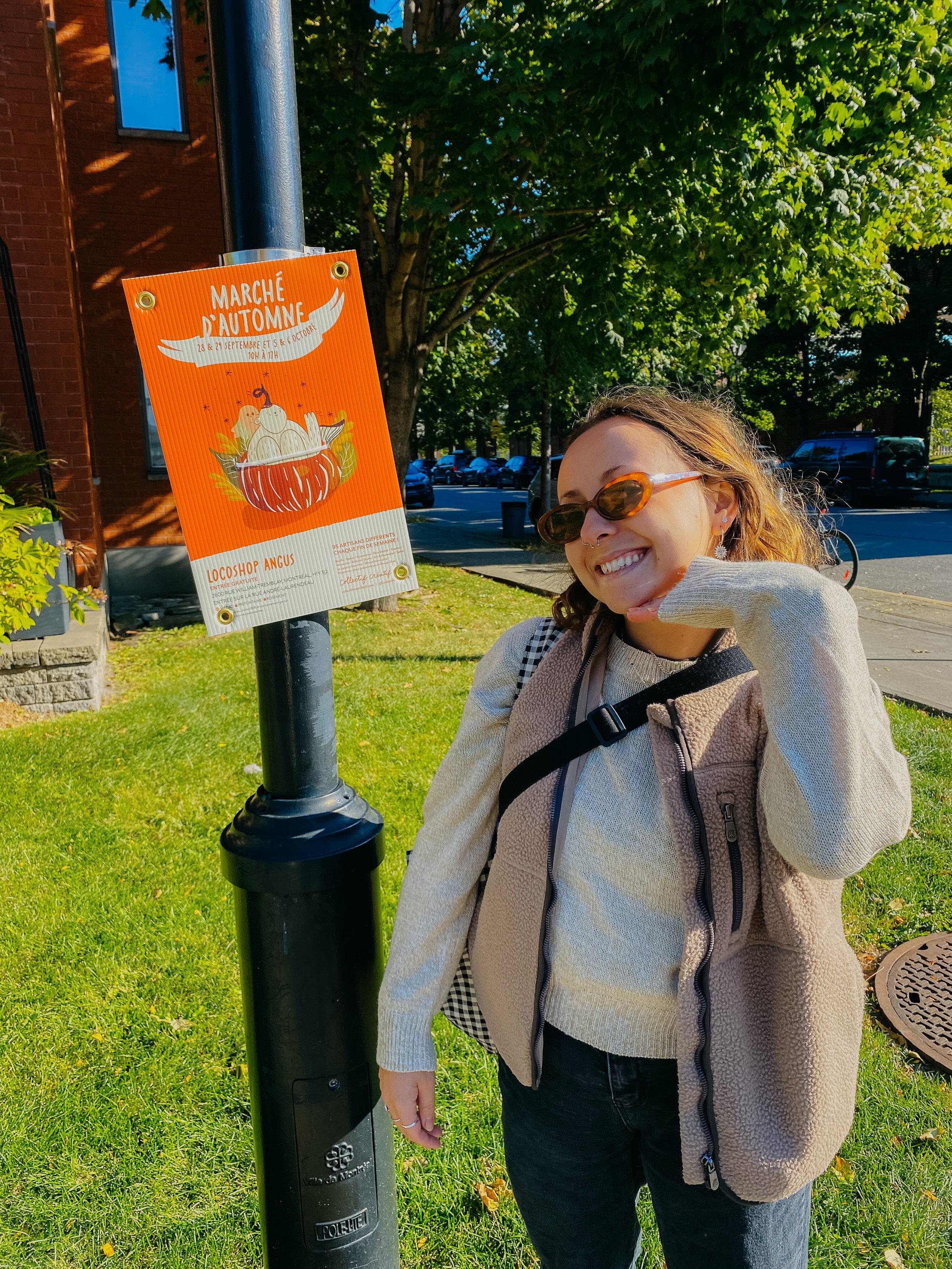 Une femme souriante avec des lunettes de soleil, un sac à dos et un pull clair, pose en plein air à côté d'un panneau annonçant un marché d'automne.
