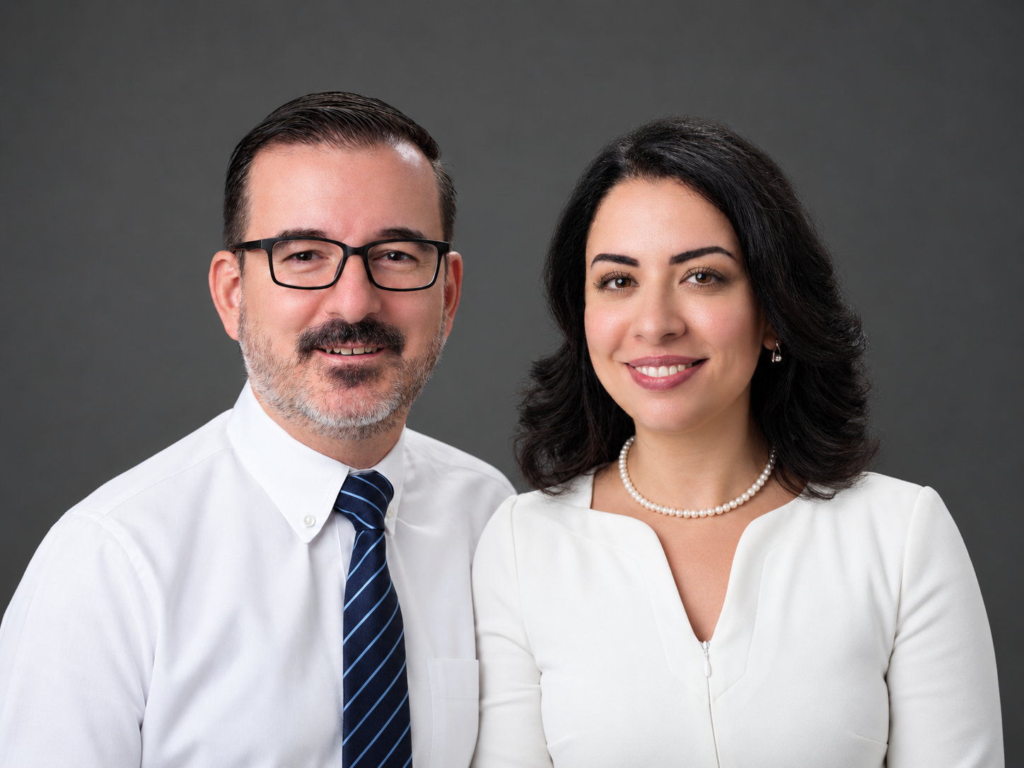A professional photo of a middle-aged man with glasses, a beard, wearing a white shirt and a striped tie, standing next to a young woman with dark hair, wearing a white dress and pearl necklace, both smiling against a gray background.