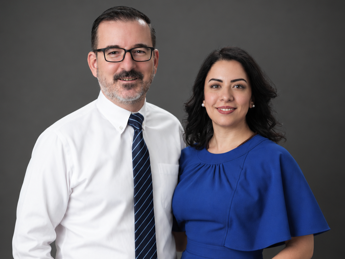 A man and woman standing together, smiling, in a studio with a dark gray background. The man is wearing a white shirt, blue striped tie, and glasses. The woman is wearing a blue dress and pearl earrings.