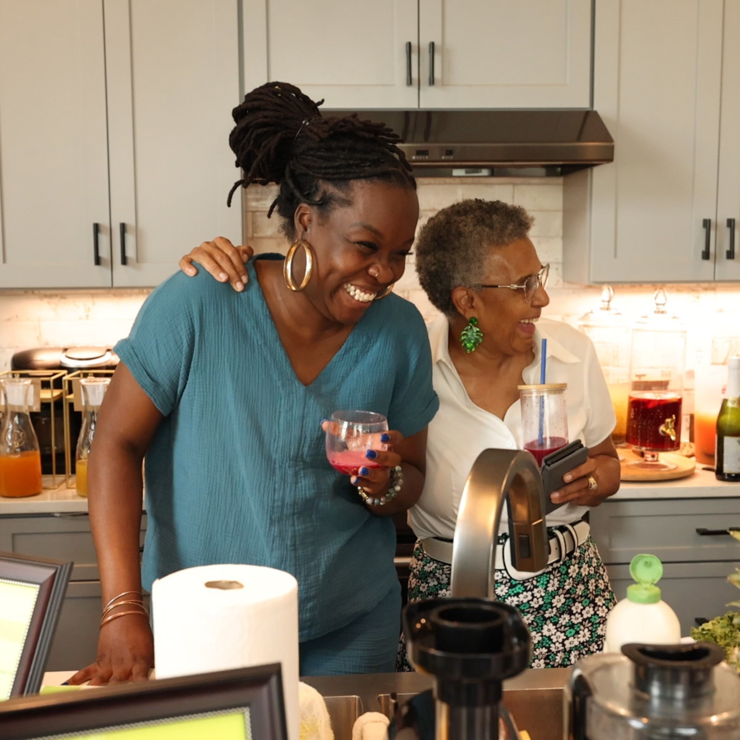 Two women are laughing and enjoying drinks together in a kitchen. One woman with dreadlocks and large hoop earrings holds a glass, while the other with glasses and green earrings holds a tumbler with a straw.