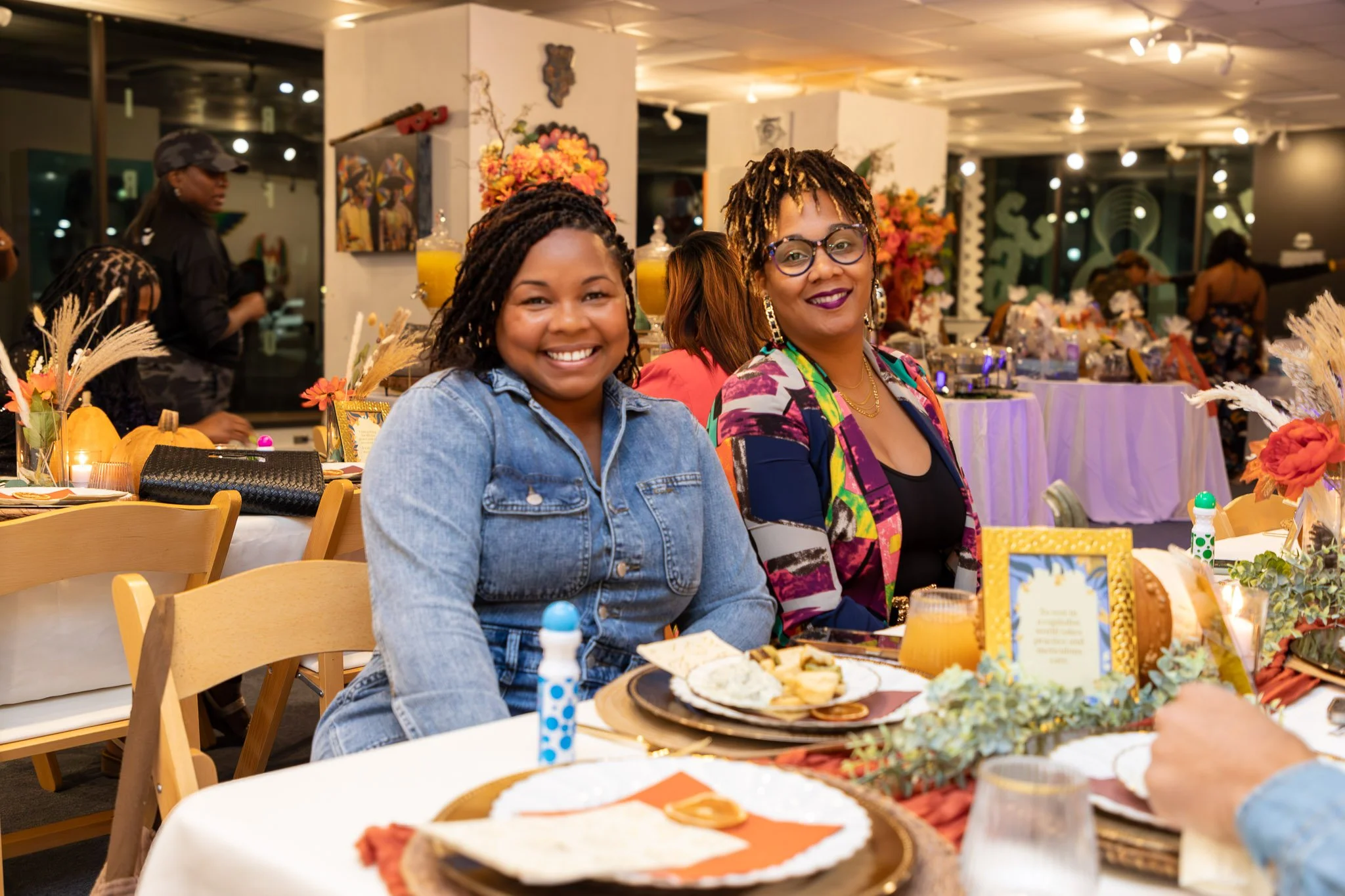 Two women smiling at a decorated indoor event. The woman on the left has curly hair, wearing a denim jacket. The woman on the right has dreadlocks, glasses, and wearing a colorful blazer. The table has festive decorations, flowers, and food.