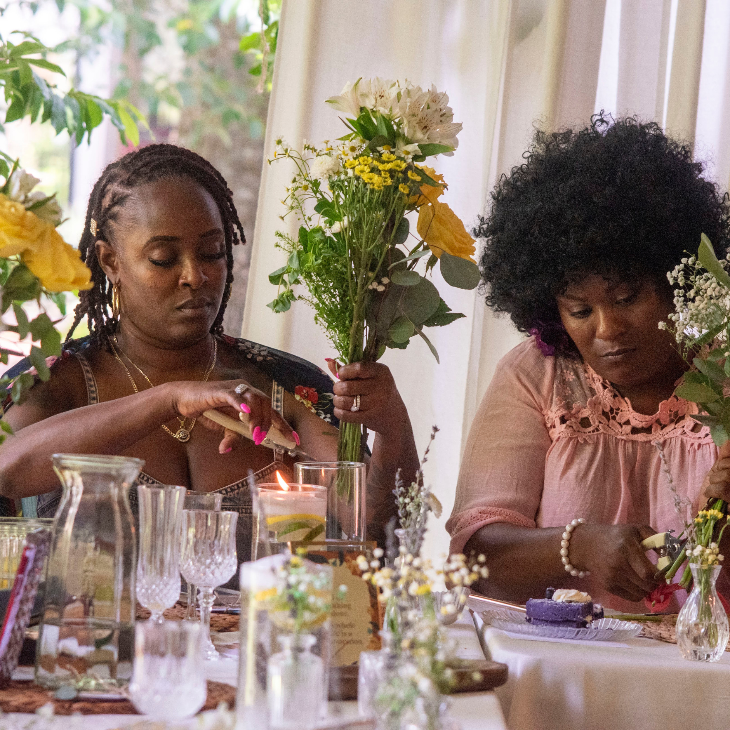 Two women arranging flowers at a decorated table, with glassware and candles, indoors near a window.