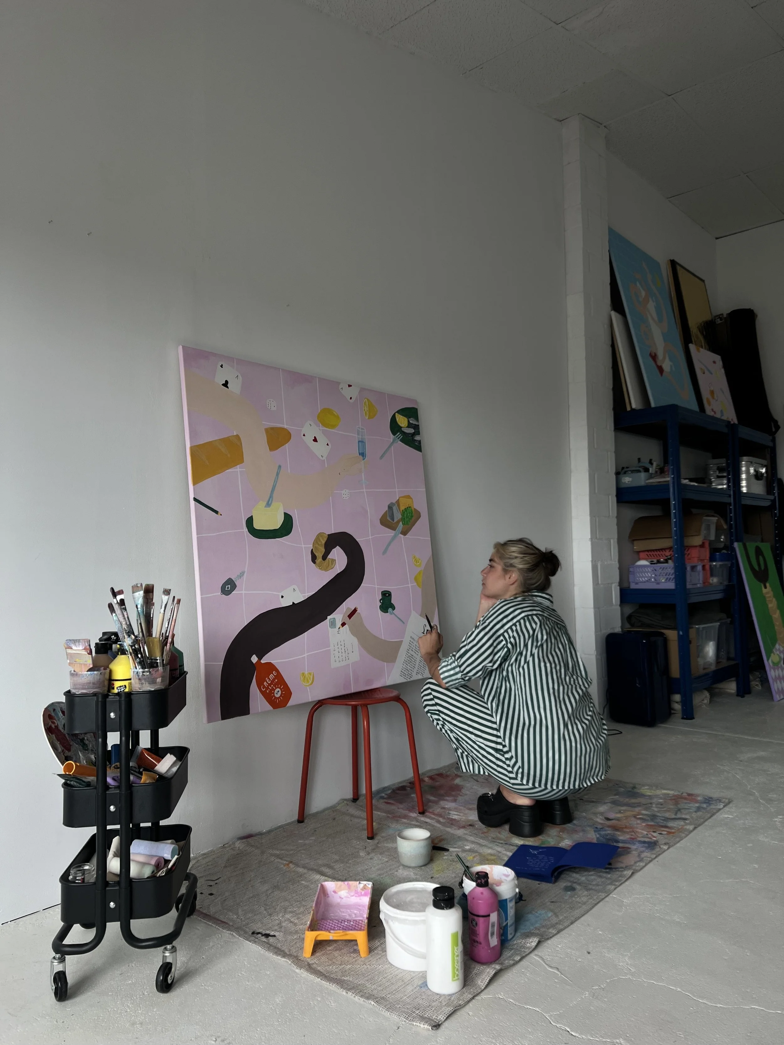 Künstlerin sitzt und malt ein großes Bild in einem Kunststudio, umgeben von Malutensilien.