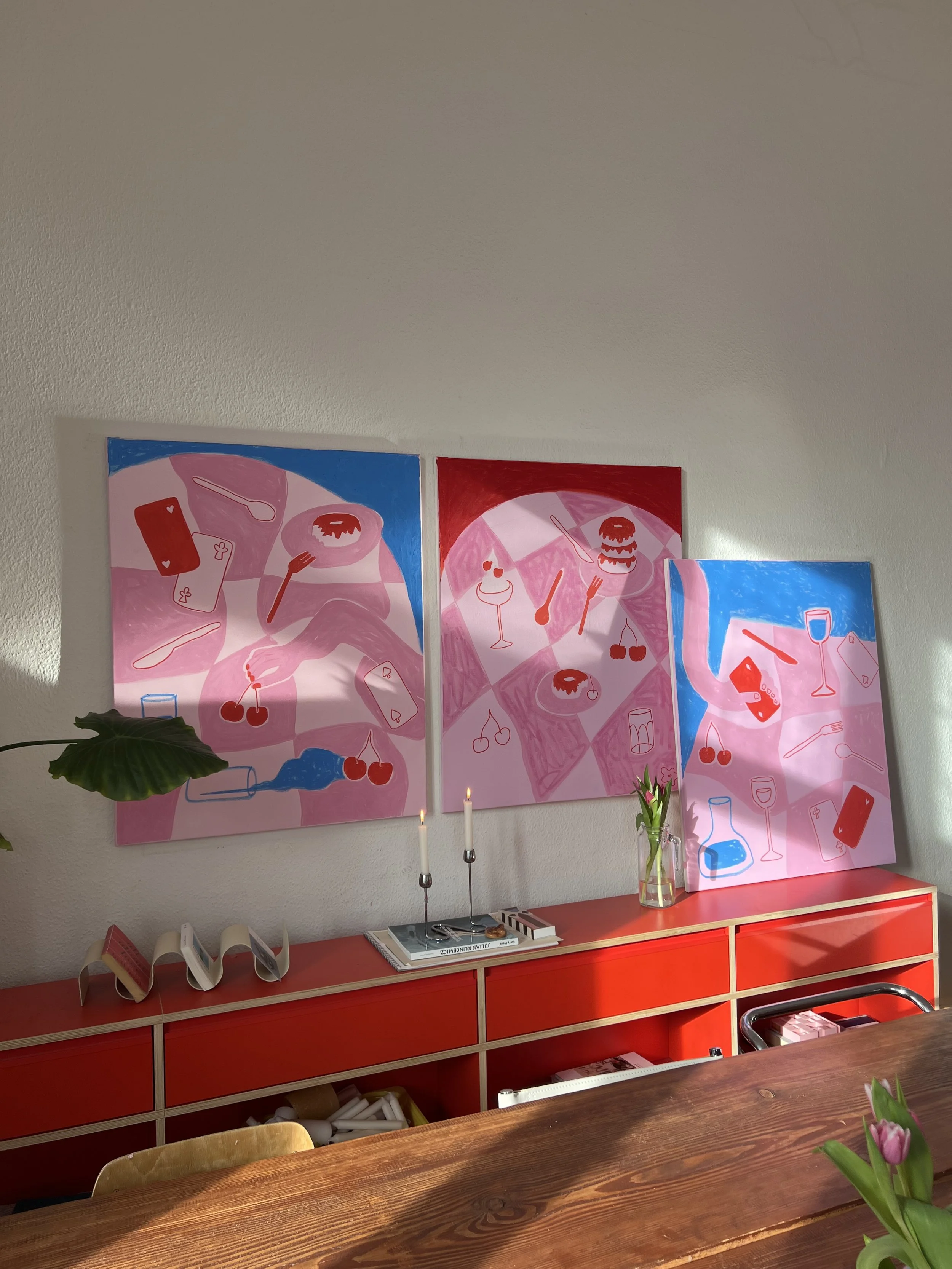 Drei bunte, abstrakte Gemälde mit Lebensmittelmotiven hängen an einer weißen Wand. Darunter steht ein rotes Sideboard mit Kerzen, Büchern, einer Vase mit Blumen und weiteren Gegenständen.