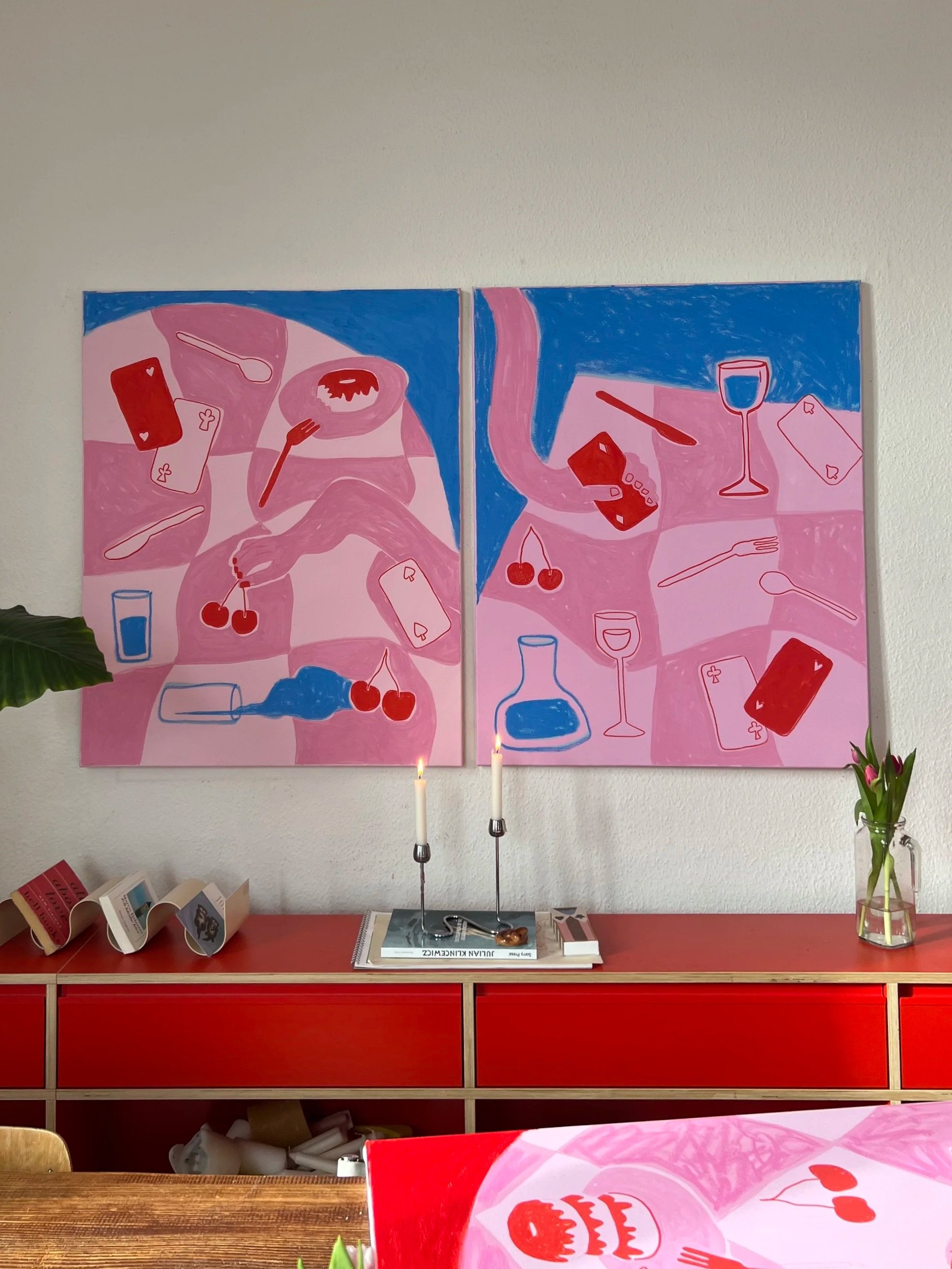 Zwei quadratische Gemälde mit pinken und blauen Farben, die Stillleben mit Flaschen, Gläsern, Karten, Früchten und Besteck darstellen. Es ist eine rote Kommode darunter, mit Kerzen, Büchern, einer Vase mit Tulpen und weiteren Gegenständen.