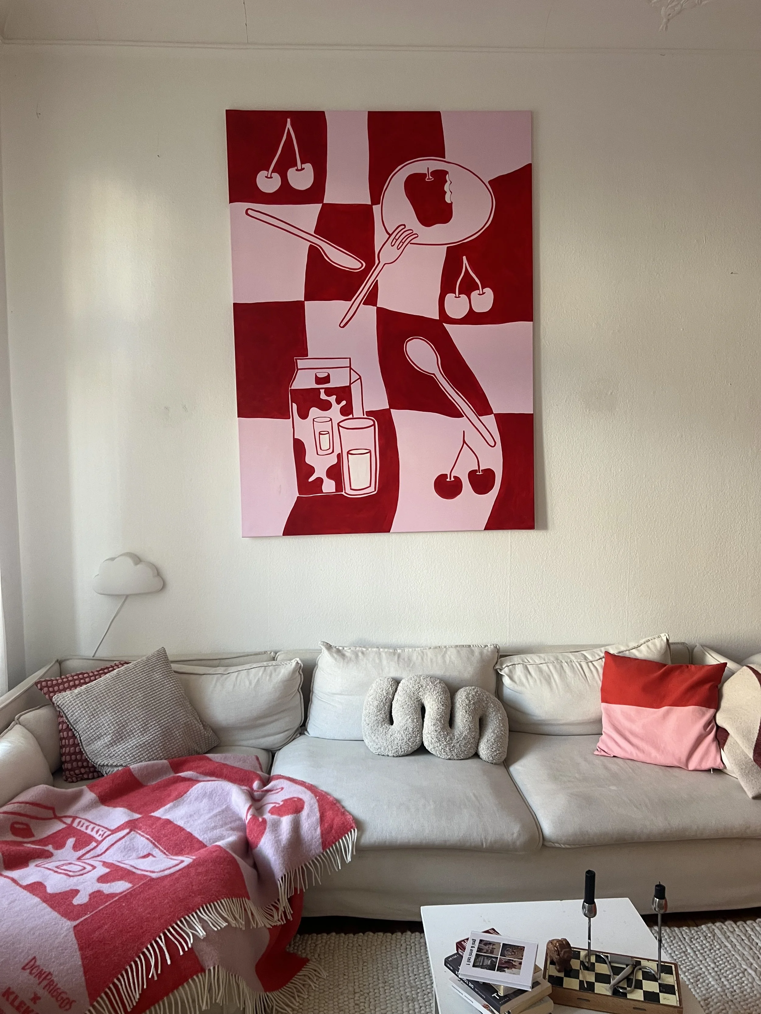 Ein Wohnzimmer mit weißen Couch, roten und grauen Kissen, einer roten und weißen Decke, einem Kunstwerk an der Wand mit roten und weißen Elementen, einem weißen Tisch mit Büchern und Dekorationen, und einer Wandlampe in Form einer Wolke.