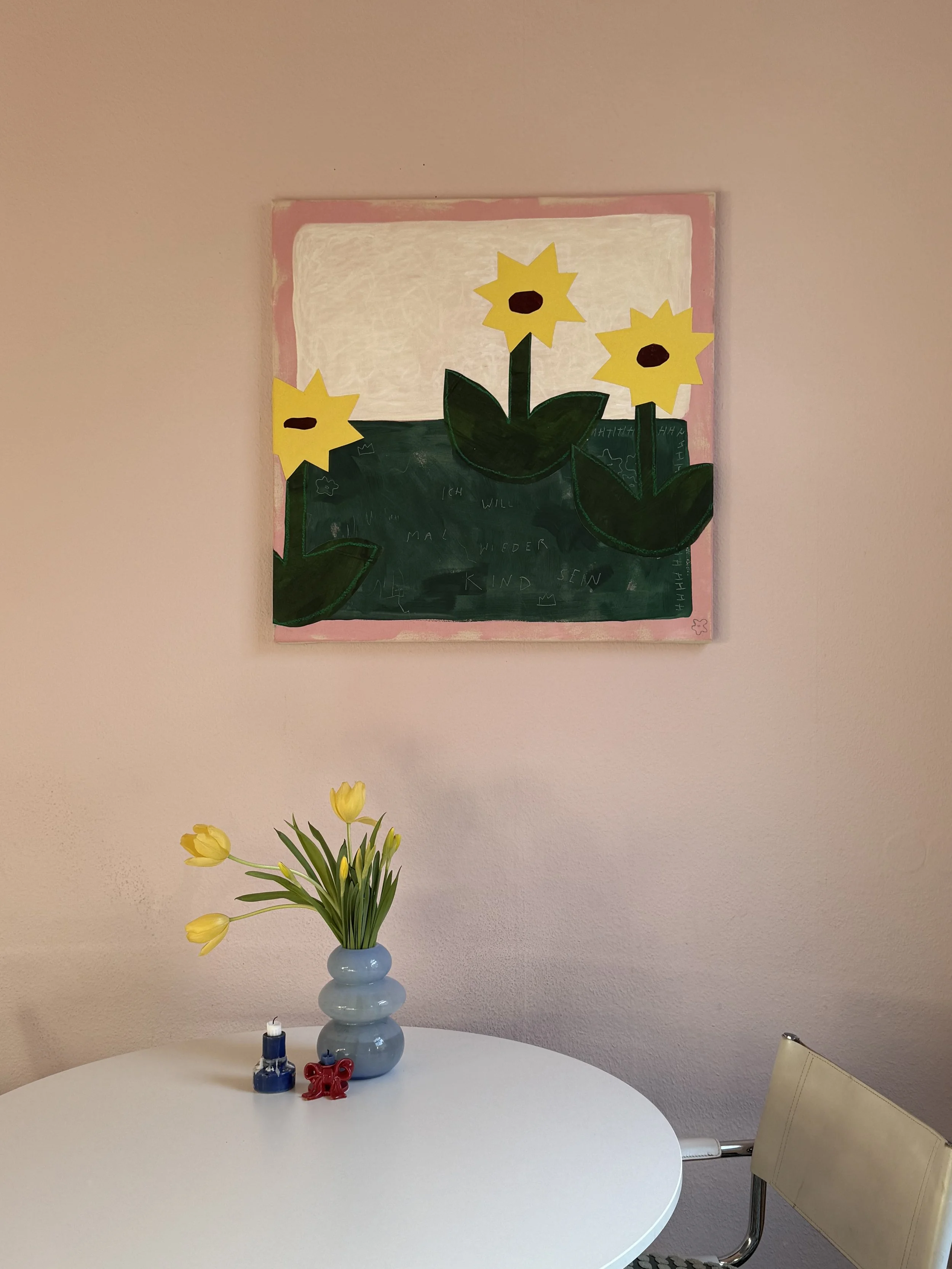 Blumenstrauß mit gelben Tulpen in einer blauen Vase auf einem weißen Tisch, hinter einem Wandbild mit Sonnenblumen und grünem Laub.