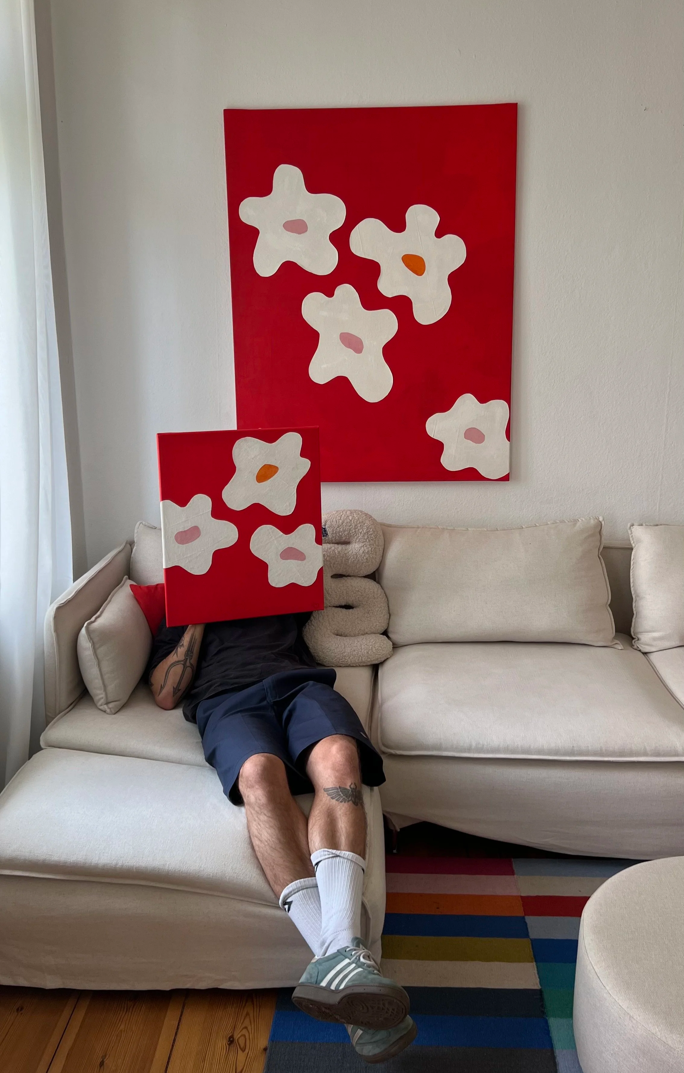 Person sitzt auf einem Sofa mit einem Kunstwerk vor dem Gesicht, das eine rote Leinwand mit weißen Blumenmotiven ist, das Kunstwerk wird auf dem Kopf gehalten.