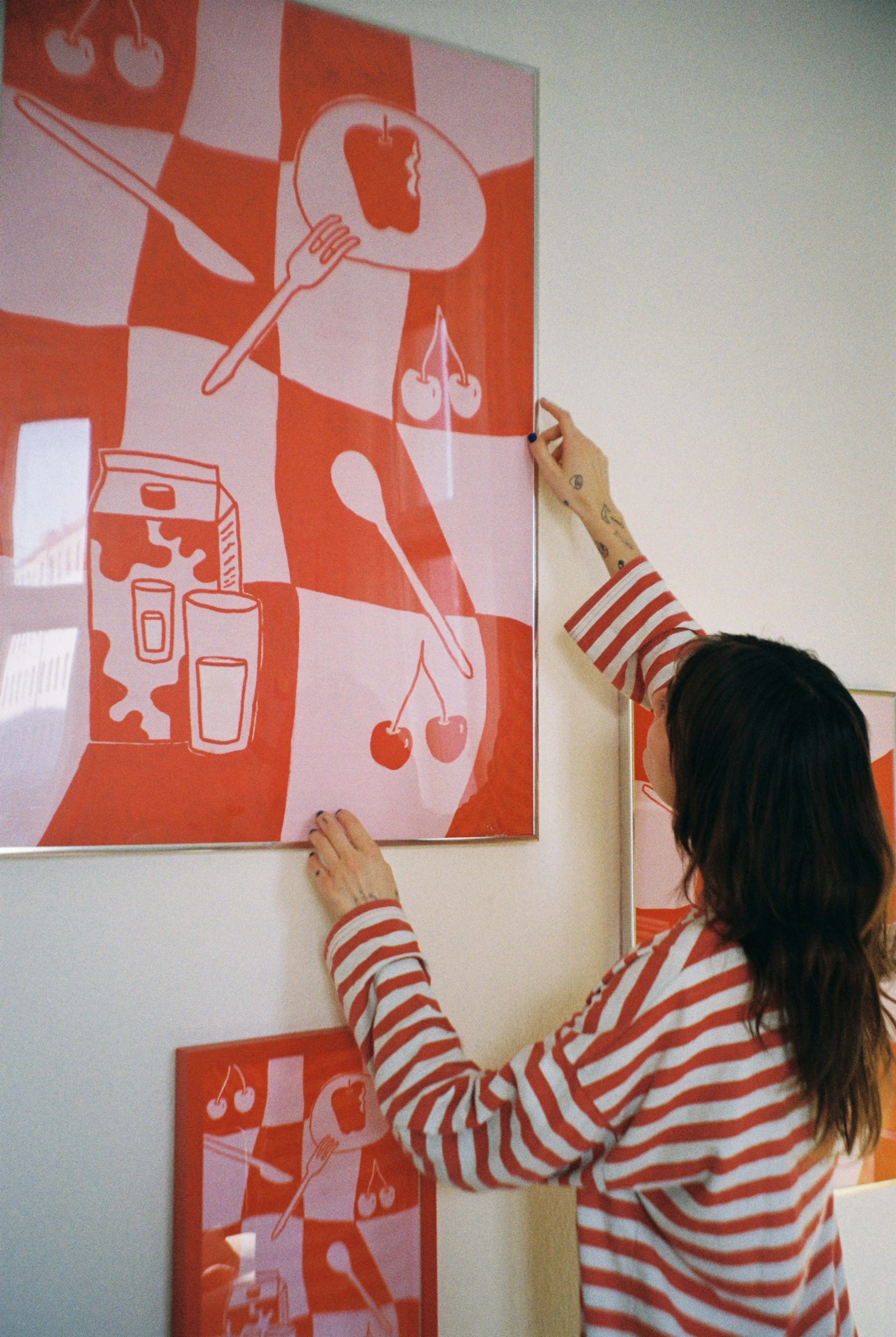 Frau, die ein Kunstwerk an einer Wand anbringt, das aus rot-weißen Kunstprints besteht, die Küchenutensilien und Lebensmittel wie Löffel, Kirschen, Joghurt und eine Paprika zeigen.