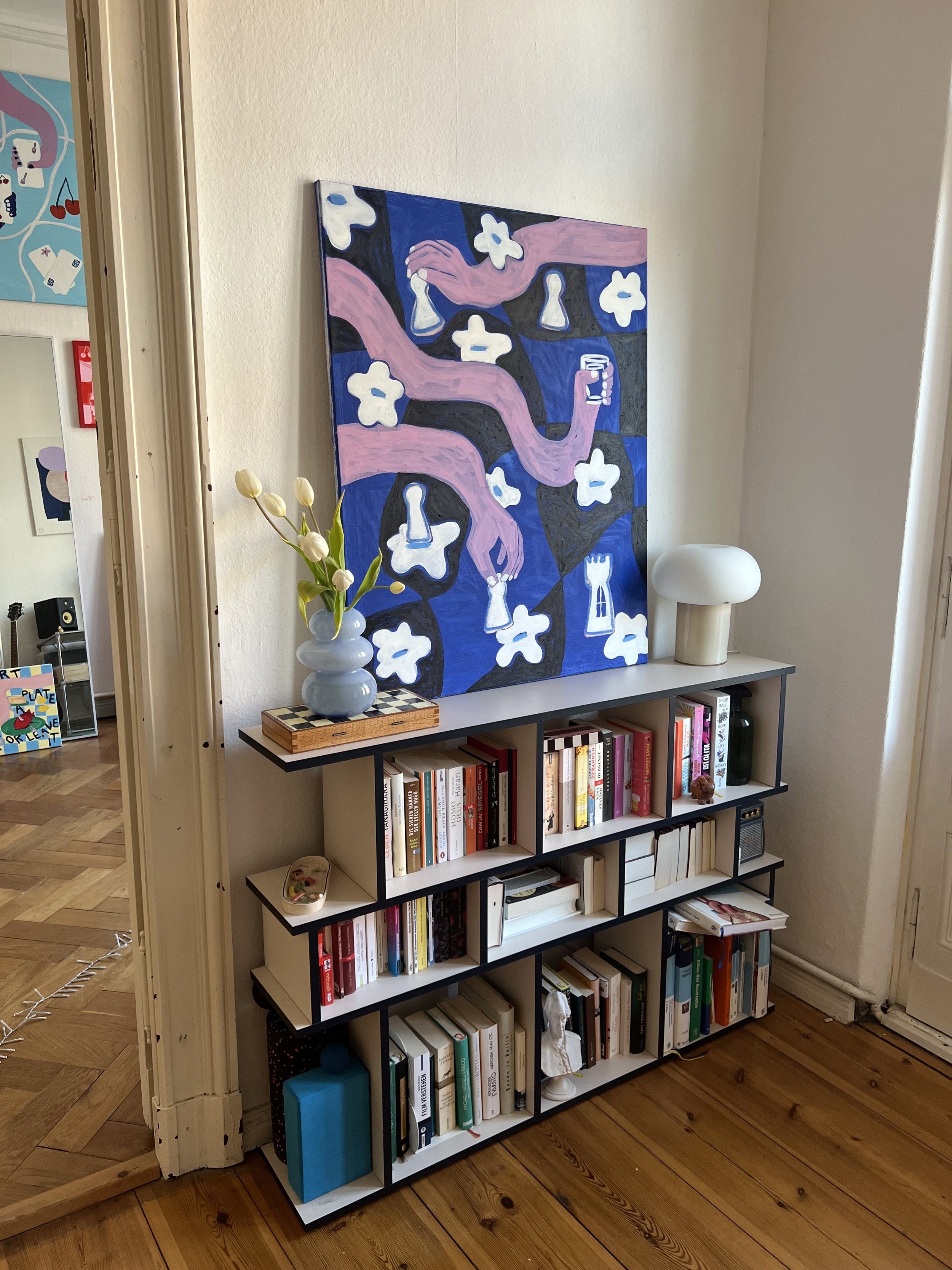 Ein Bücherregal mit mehreren Regalböden, einer Vase mit weißen Tulpen, einem weißen Lampenschirm und einem Gemälde mit pinken, blauen und weißen Elementen gegen die Wand.