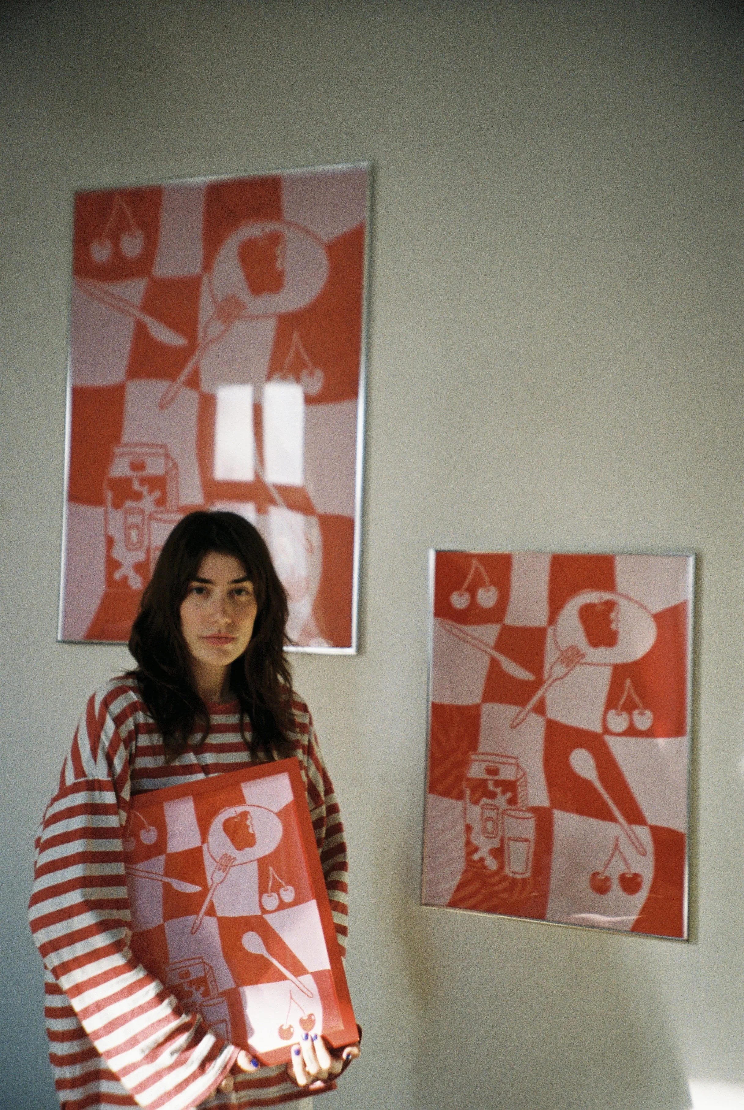 Eine junge Frau mit braunen, gewellten Haaren trägt ein rotes und weiß gestreiftes Hemd. Sie hält ein rotes Bild mit einer grafischen Illustration in den Händen. Im Hintergrund sind zwei große, gleich gestaltete Bilder an der Wand, die ebenfalls die 