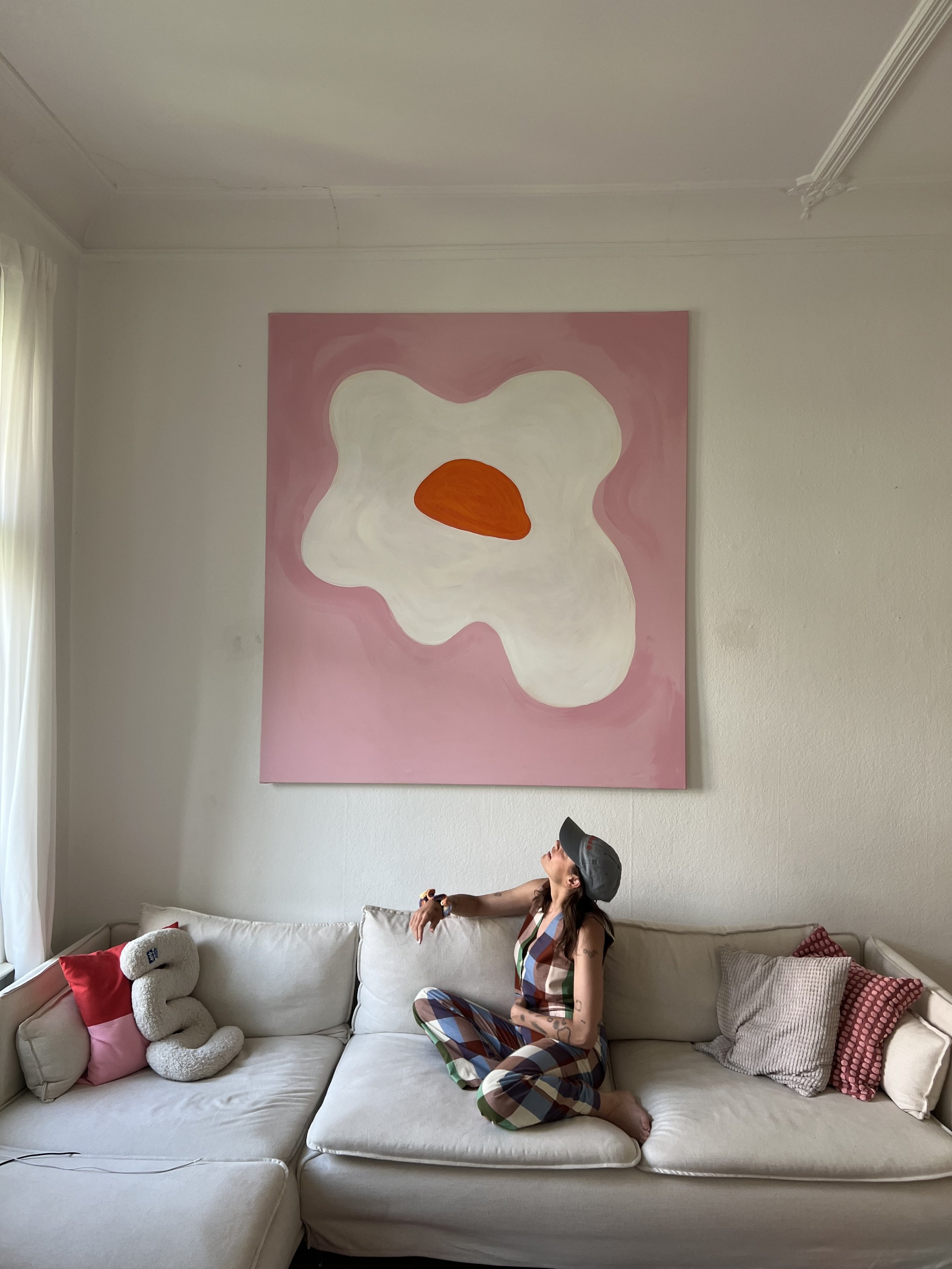 Frau sitzt auf einer weißen Couch, trägt bunte Karotten-Overall und eine Mütze, neben einem Plüschtier in Form einer Zahnpasta, im Wohnzimmer mit pastellfarbenen Kissen und einem großen abstrakten Gemälde an der Wand.