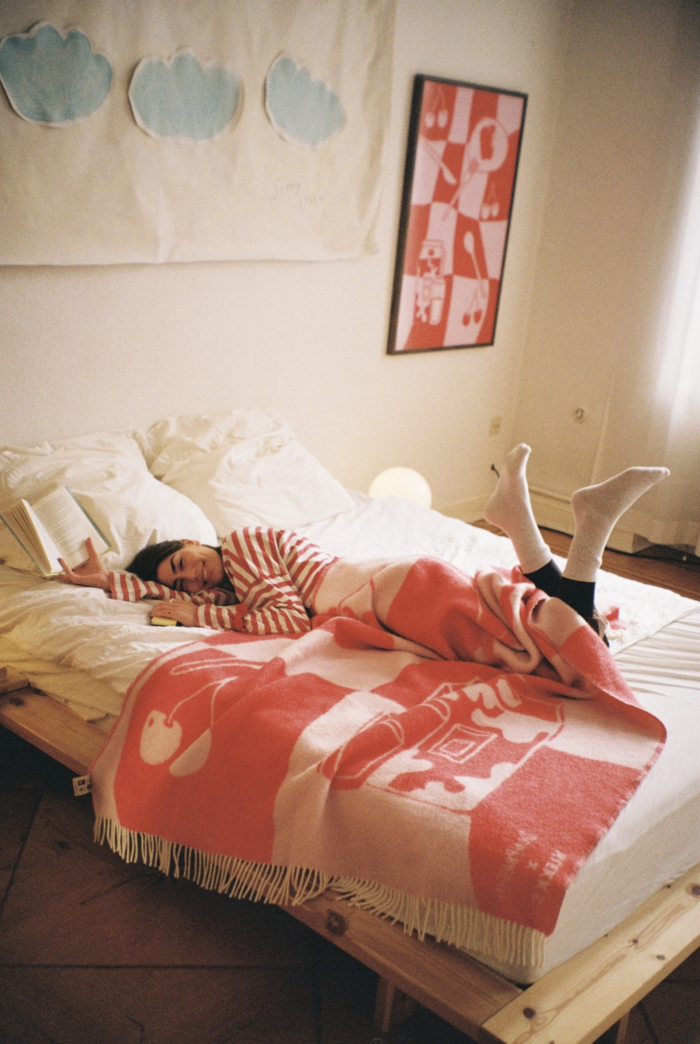 Jugendliche Frau liegt entspannt auf einem weißen Bett mit einem Buch in der Hand, ist in einer roten und weißen Strickjacke und Leggings gekleidet, und hat die Beine in die Luft gestreckt, um sich auszuruhen. Der Raum ist gemütlich mit Wanddekoration und einem weichen, roten Decke.