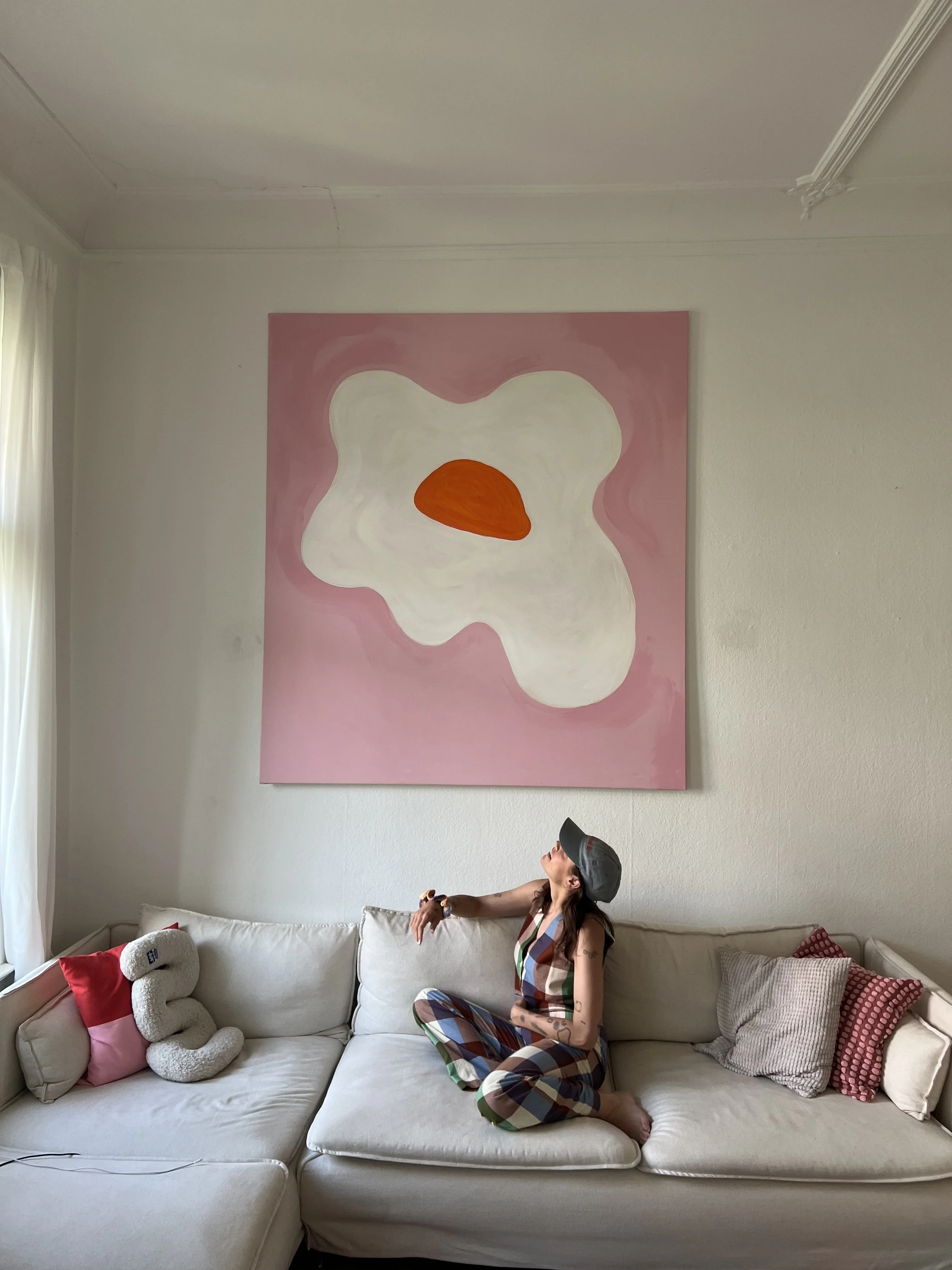 Eine Frau sitzt in einem weißen Sofa vor einer großen abstrakten Wandmalerei mit weißen, pinken und braunen Farben. Sie trägt eine bunte Jumpsuit und eine graue Cap.