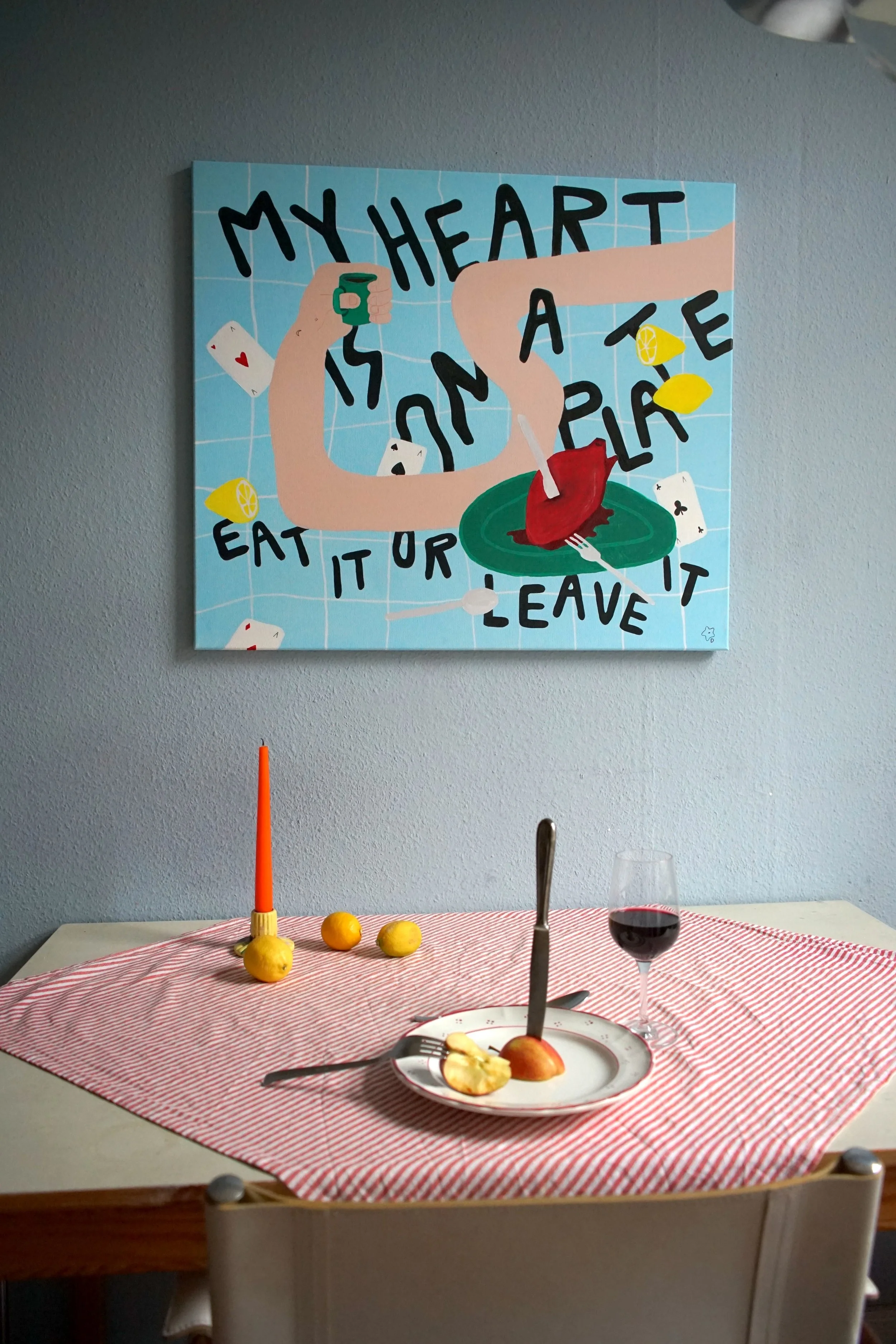 Innenaufnahme eines Tisches mit rot-weiß karierter Tischdecke, einem Teller mit Apfel, Messer, halbierter Frucht, einem Weinglas mit Rotwein, drei Zitronen, einer roten Kerze, und einem Wandbild mit einem menschlichen Arm, der einen grünen Fisch hält, und schwarzen Text.