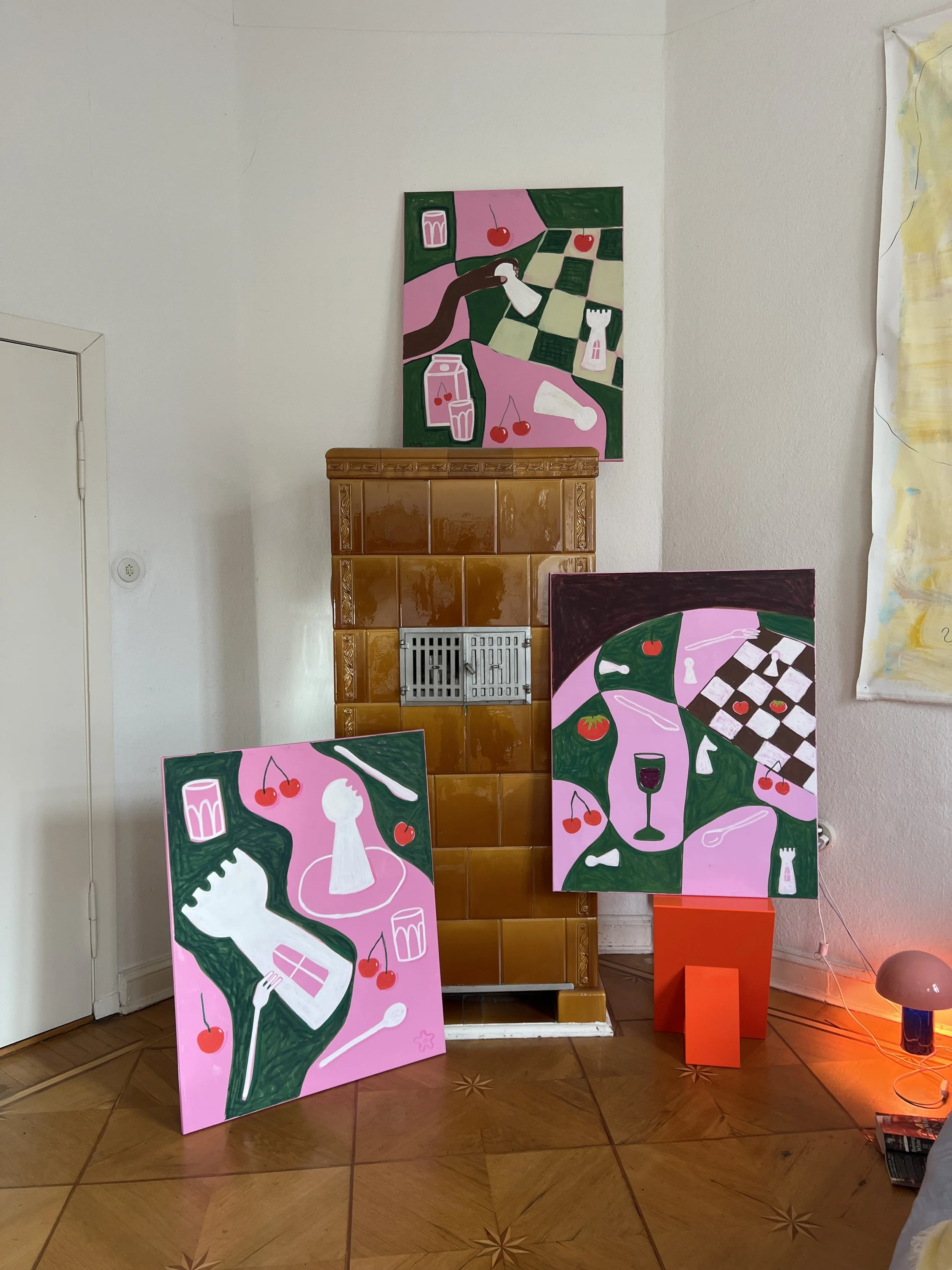 Vier bunte Gemälde mit pinkem Hintergrund, die verschiedenen Gegenstände wie Kirschen, Gläser, Besteck, und Schachfelder darstellen, sind in einem Raum aufgestellt.