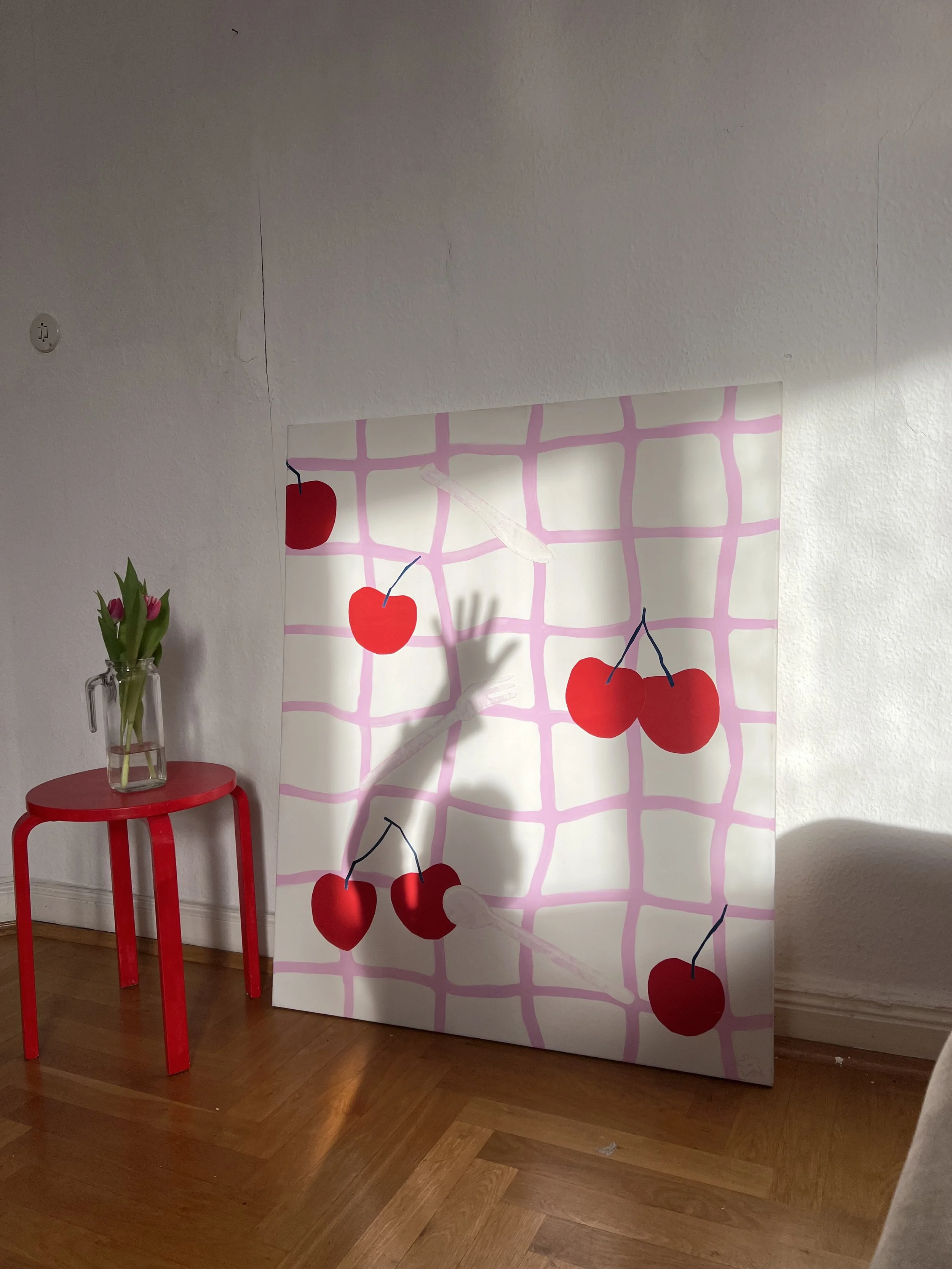 Ein bemaltes Bild mit roten Kirschen auf einem weißen Hintergrund mit pinkem Gitternetzmuster, das gegen eine weiße Wand lehnt, neben einem roten Spieltisch mit einer Vase mit Tulpen.