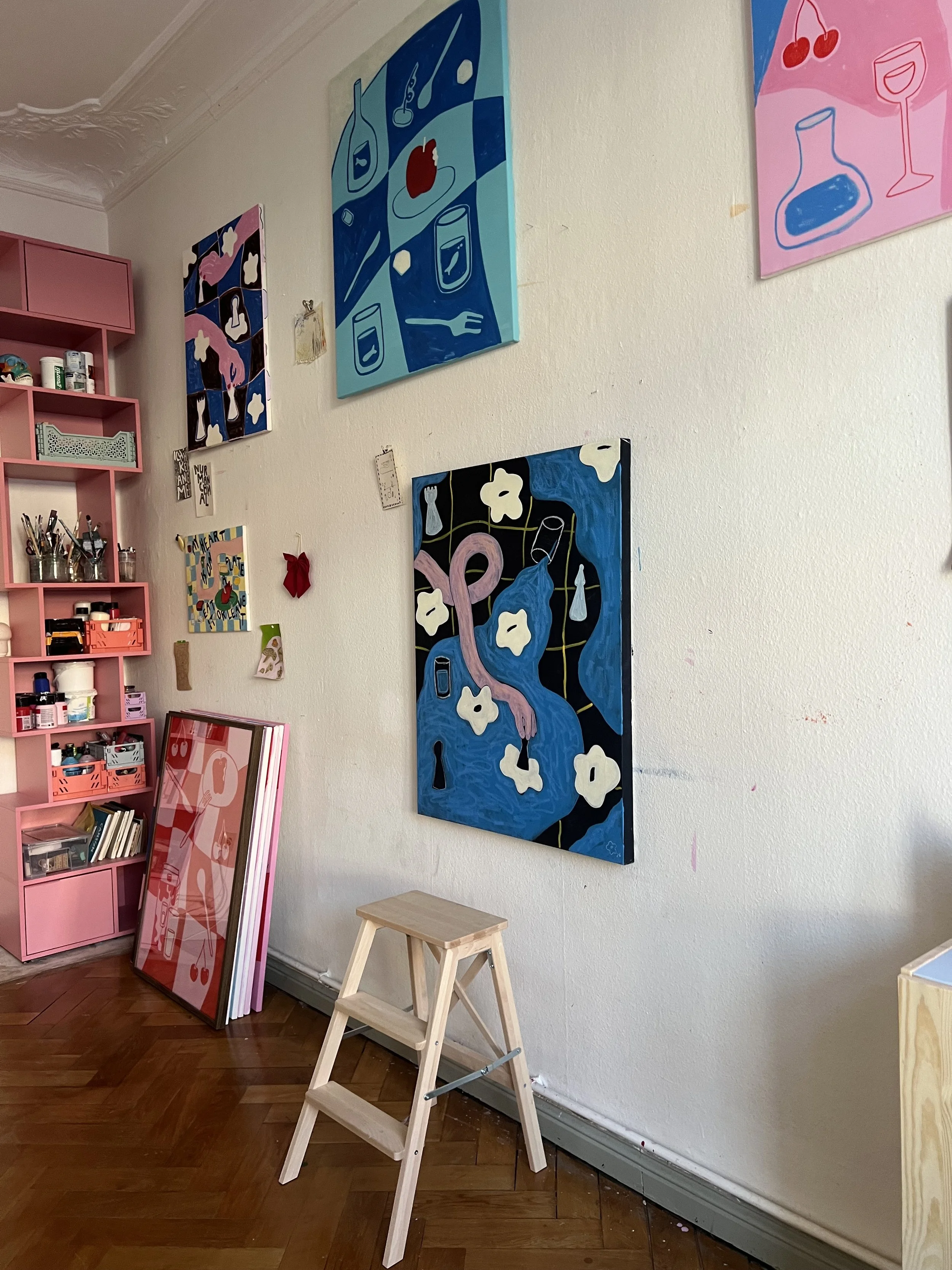Bunte Kunstwerke hängen an einer weißen Wand, darunter steht ein kleiner Holhocker. Ein pinker Regal mit verschiedenen Gegenständen steht rechts neben den Kunstwerken.