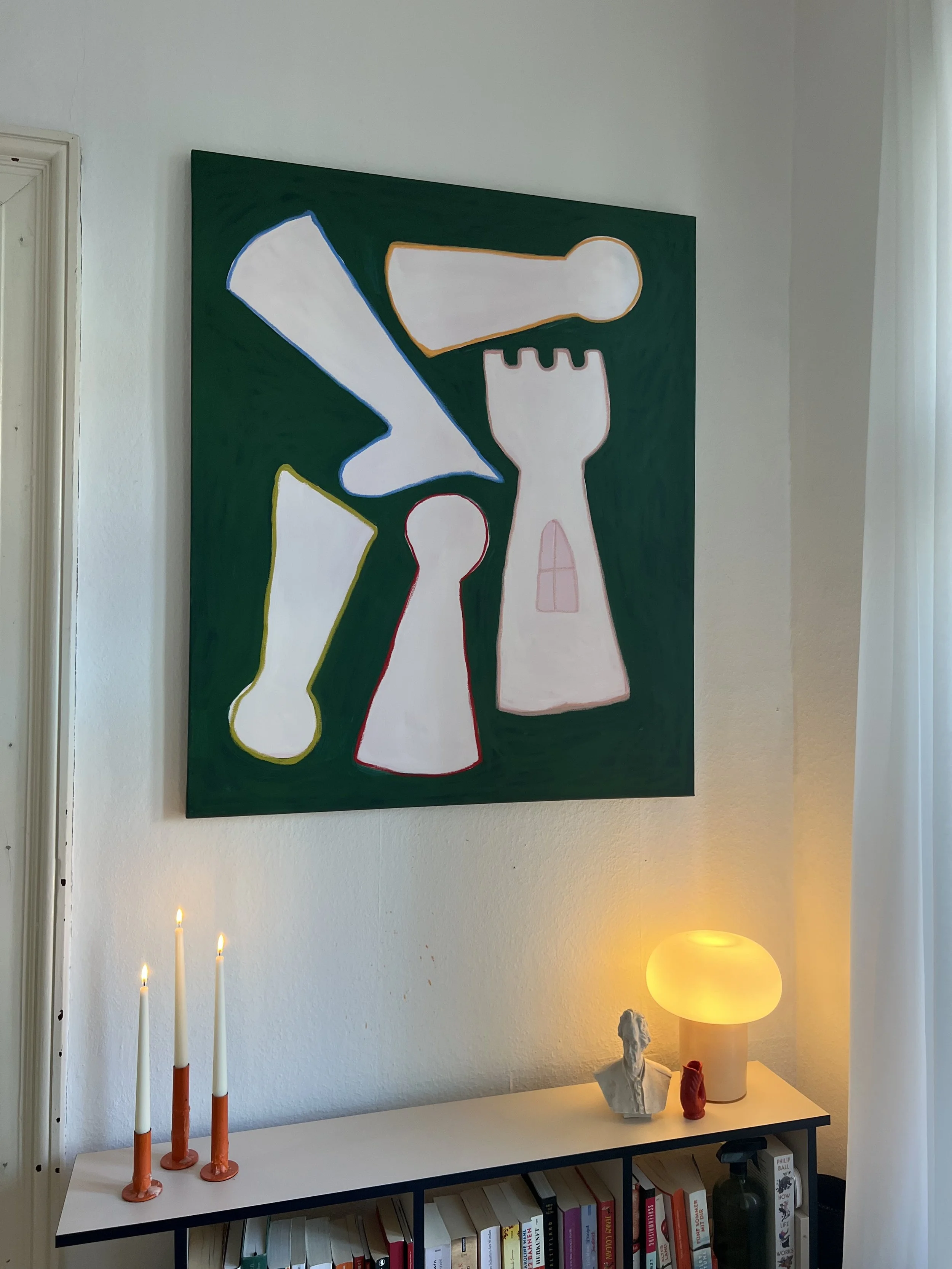 Ein Wandbild mit abstrakten Formen in verschiedenen Farben, darunter Weiß, Blau, Rot, Pink und Orange, hängt an einer weißen Wand. Darunter steht ein weißer Tisch mit Kerzen, einer Skulptur und einem Lampen in einer gemütlichen Ecke.