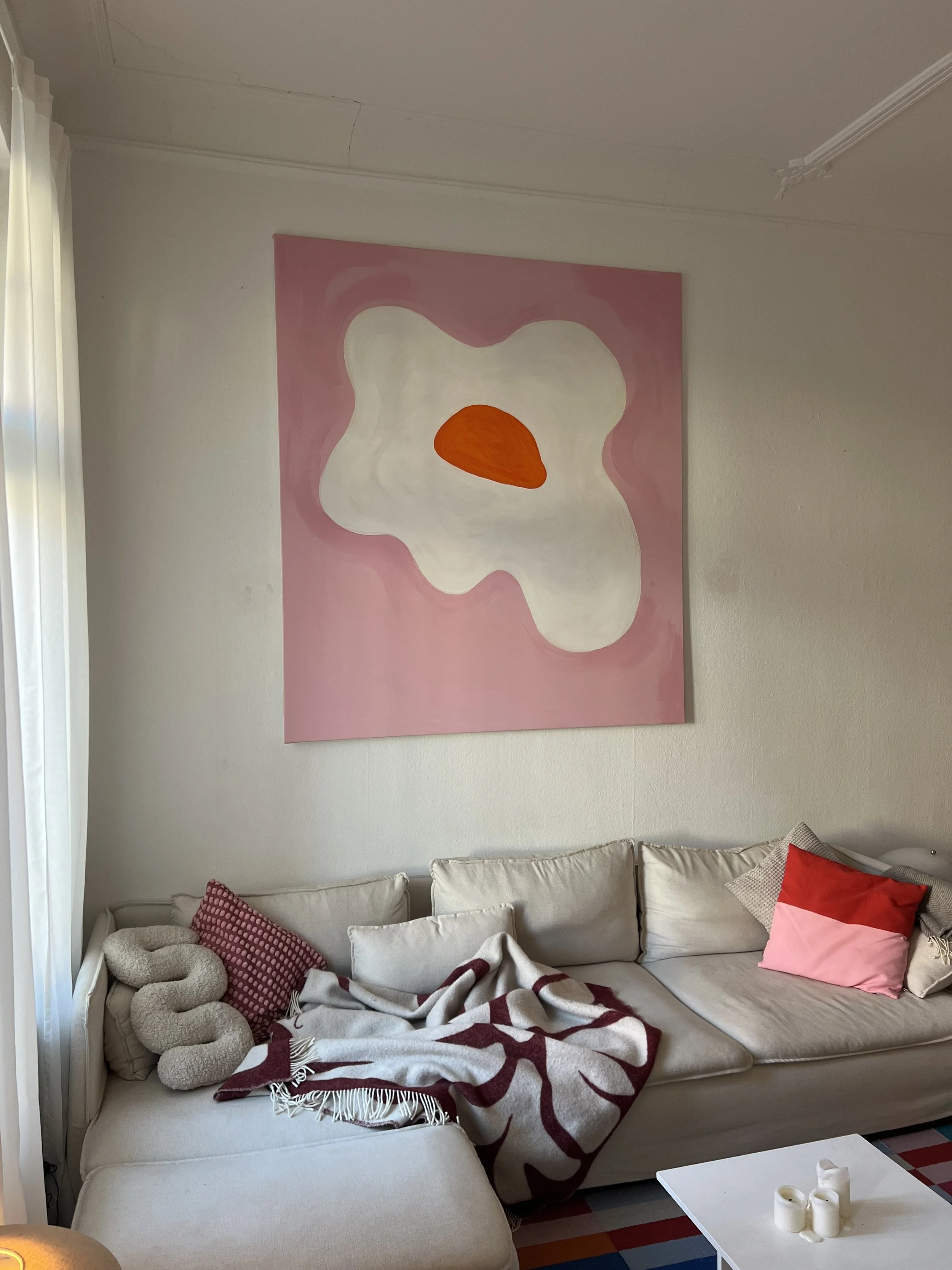 Ein weißes Sofa mit verschiedenen Kissen und einer Decke, darüber ein Gemälde mit abstrakter Form in Weiß und Orange auf rosa Hintergrund, im Raum mit weißen Wänden und Vorhängen