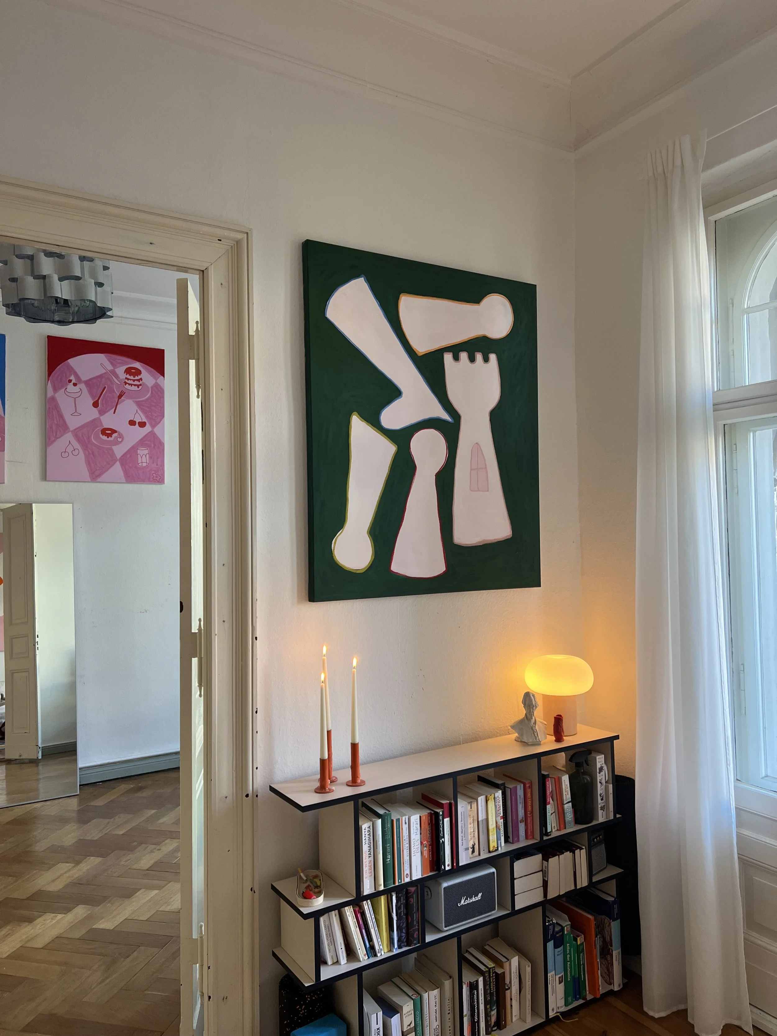 Ein Wohnzimmer mit einer großen, abstrakten grün-weißen Malerei an der Wand, Bücherregal mit Büchern und Dekorationsgegenständen, zwei brennende Kerzen, einer kleinen Skulptur, einem Lichtobjekt und einem Lautsprecher auf dem Regal, helle Wände, eine große Fensterseite mit weißen Vorhängen.