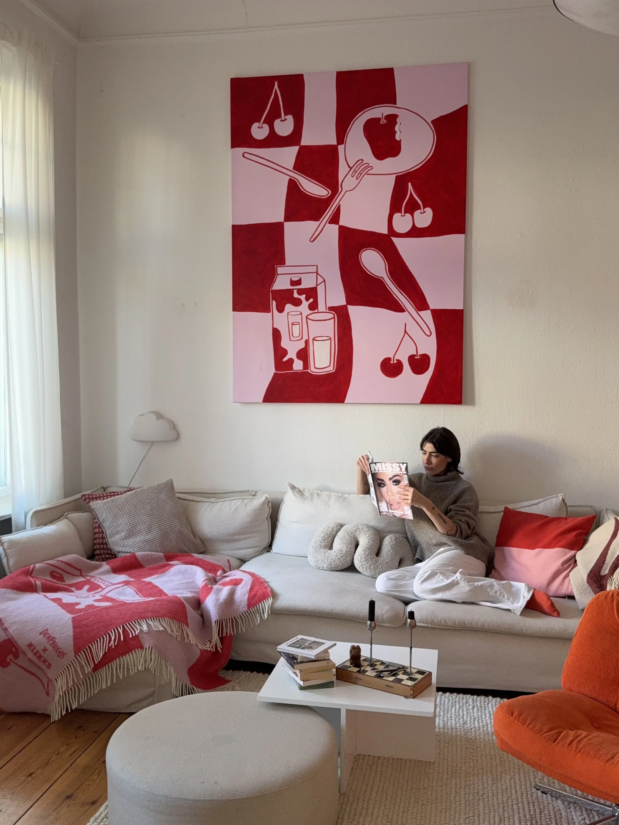 Ein Wohnzimmer mit einer weißen Couch, auf der eine Frau sitzt, die eine Zeitschrift liest. An der Wand hängt ein großes, abstraktes Kunstwerk in Rot- und Pinktönen. Der Raum ist gemütlich mit Kissen, einer roten Decke und einem kleinen Beistelltisch mit Büchern und Kerzen. Es gibt ein Fenster mit weißen Vorhängen und einen orangefarbenen Sessel.