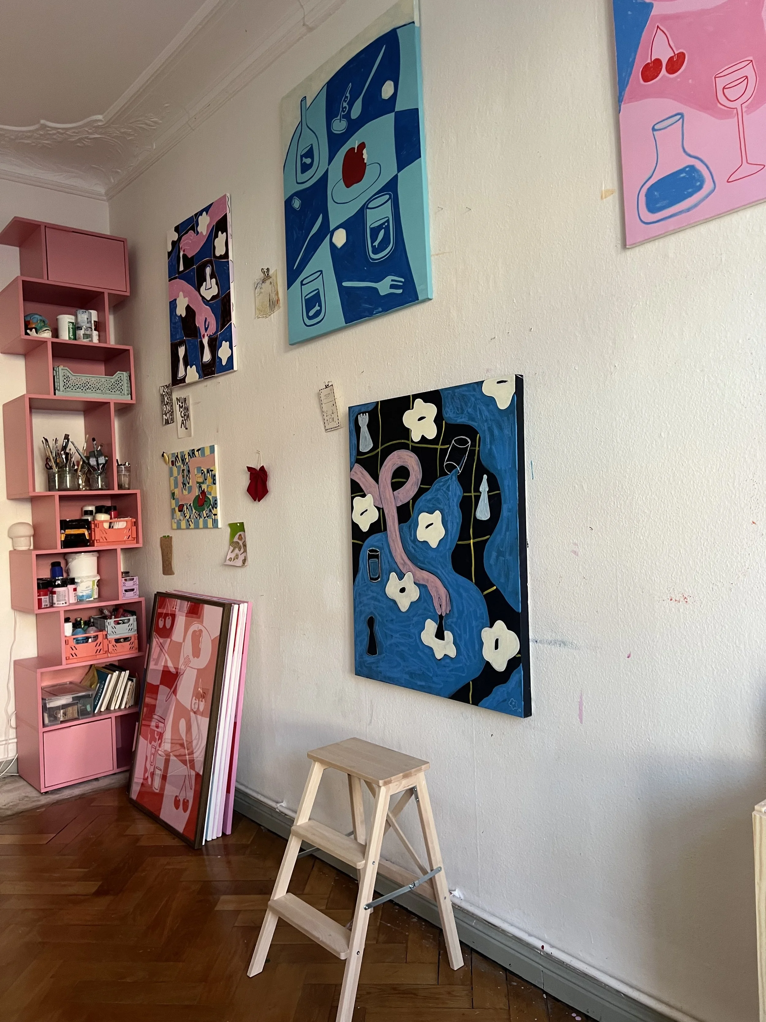 Kunstwerke an einer weißen Wand, darunter ein Holzleiter, auf einem Holzboden, mit pinkem Regal auf der linken Seite.