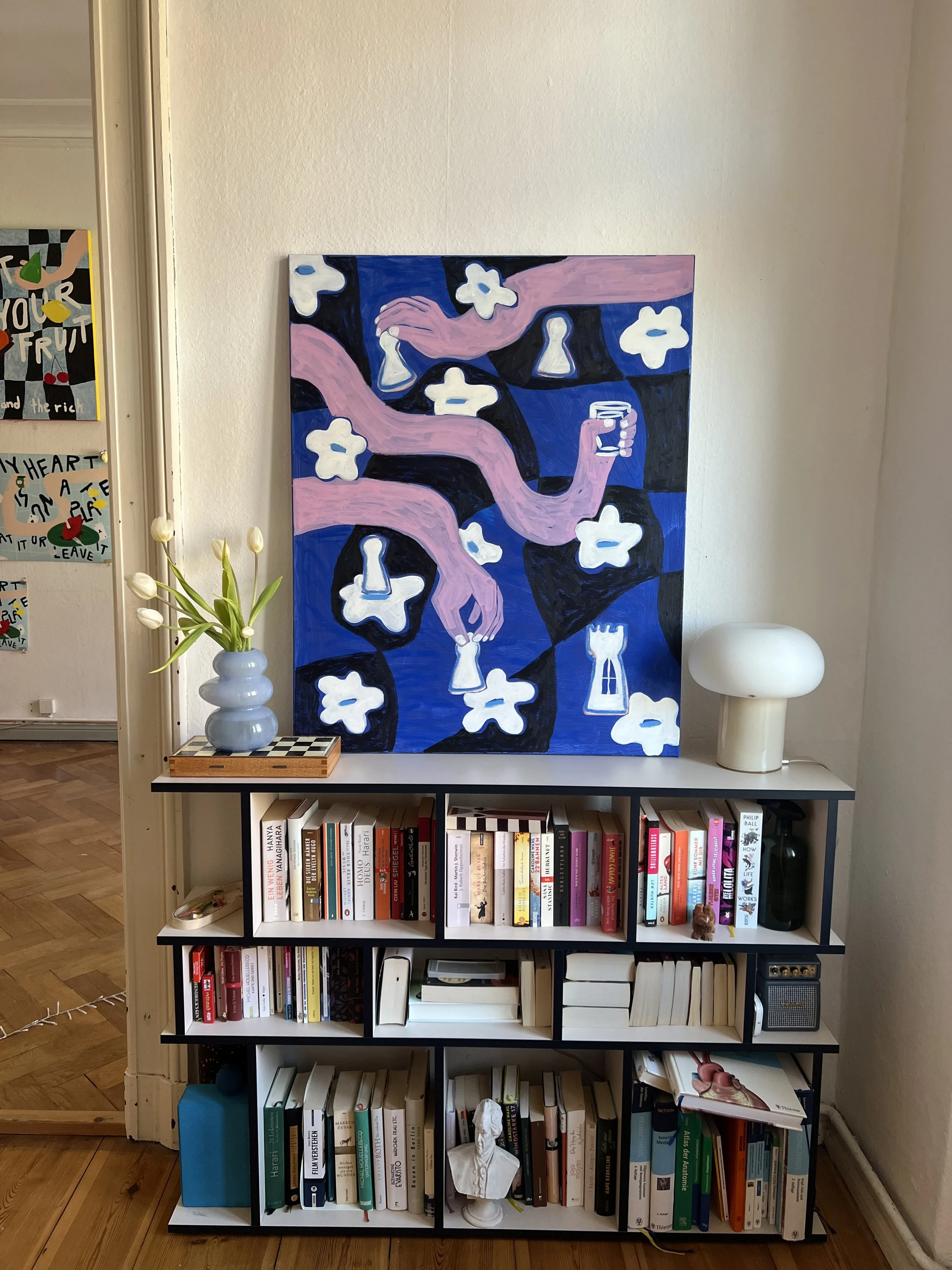 Ein Bücherregal mit mehreren Fächern, auf einem Farbfoto im Hintergrund eine blaue Wand mit einem Gemälde mit pinken Tentakeln, weißen Blumenformen, Schachbrettmuster und Schlosssymbolen, links daneben eine Vase mit weißen Tulpen.