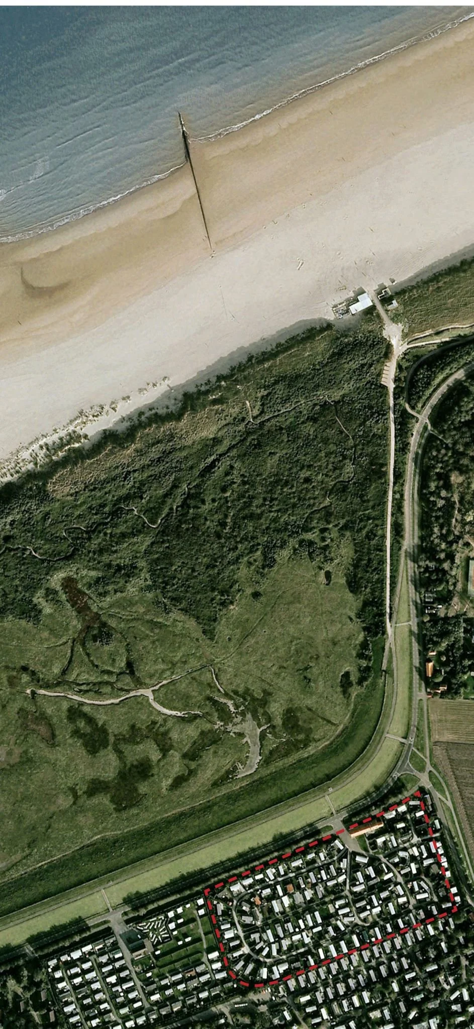 Luchtfoto van een kustlijn met strand, zee en een bosgebied ernaast, en het vakantiepark met autowegen onderaan de afbeelding.