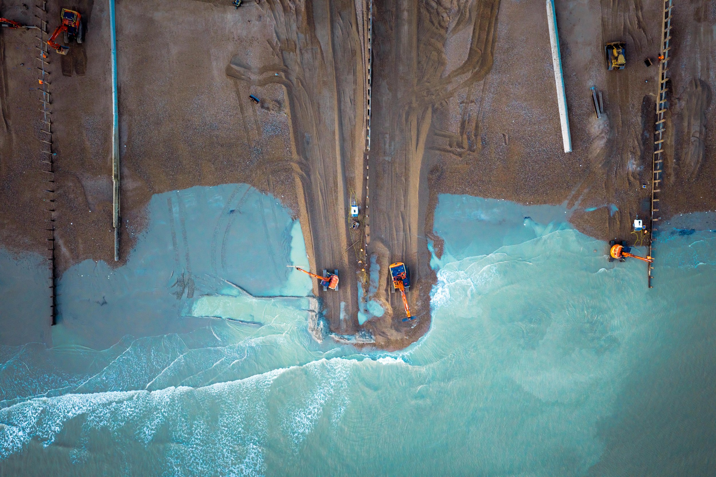 Aerial view of a construction site on a beach in Hove with machinery working on the shoreline, excavating and moving sand, with ocean waves crashing onto the shore.