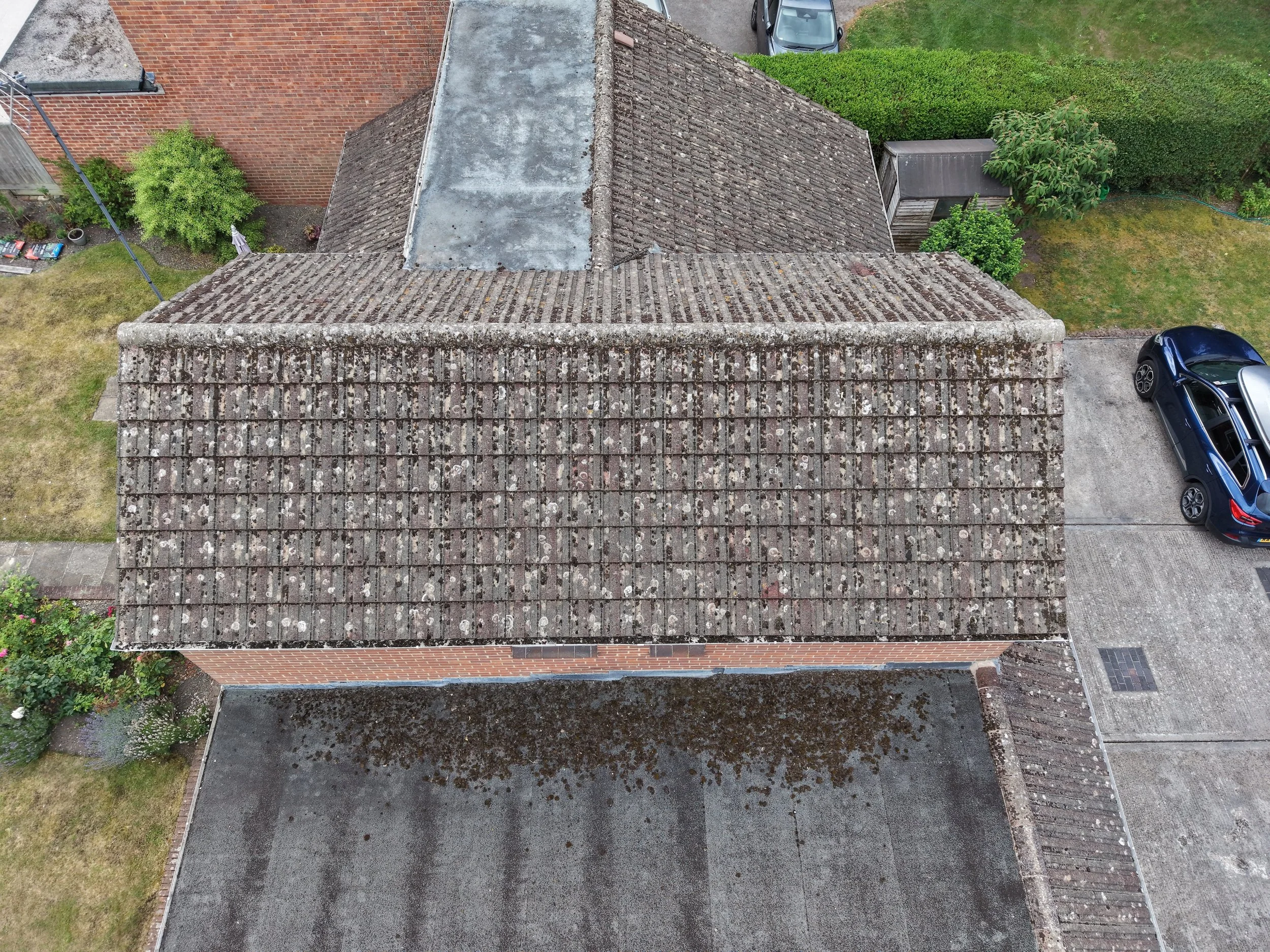 Overhead view of rooftops with brown tiles, one with moss and lichen, next to a gray flat surface, green garden, and parked cars.