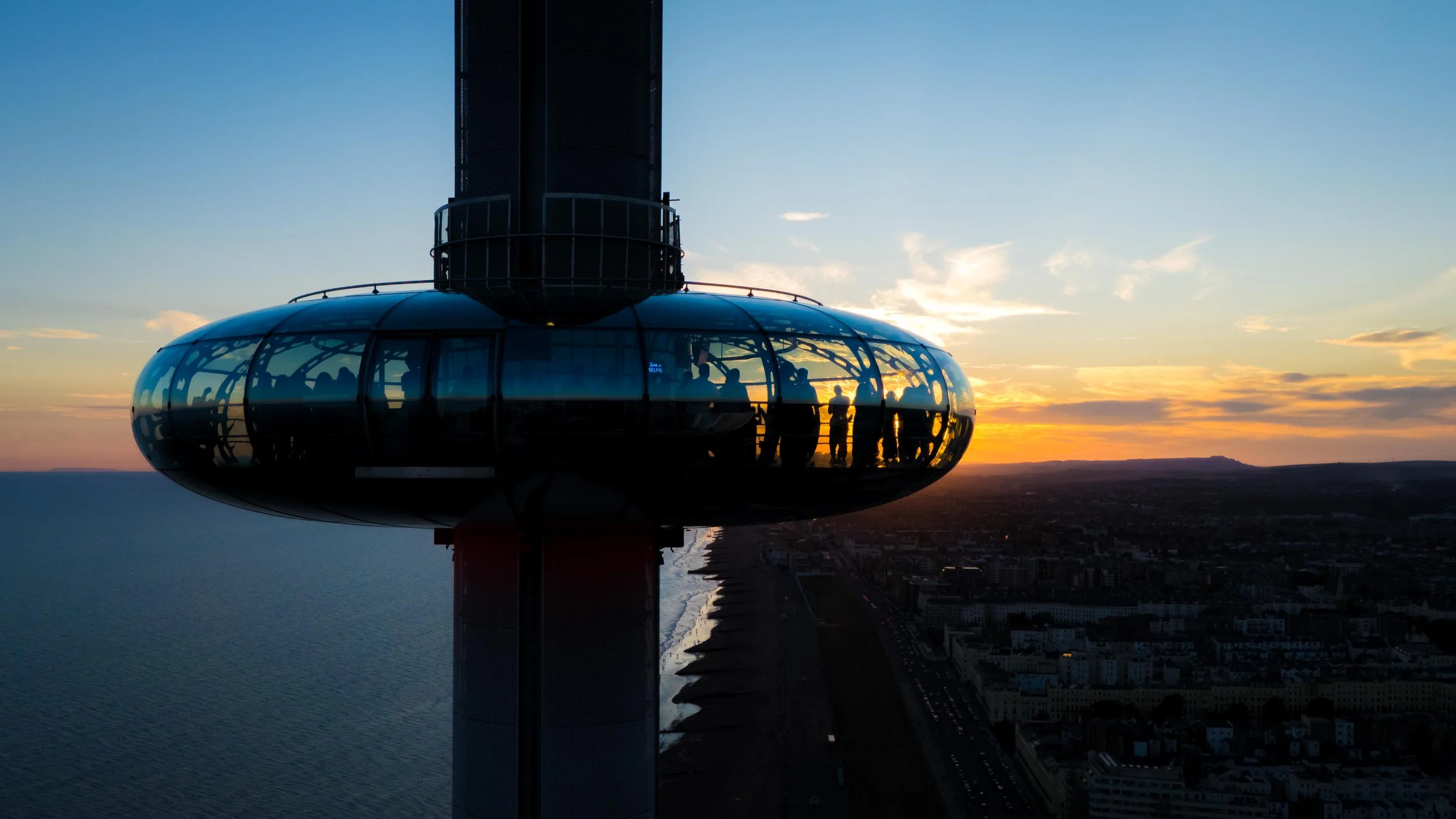 Brighton i360 observation tower with a rounded glass-enclosed viewing area, silhouetted against a sunset sky, with people visible inside the glass. The background includes a coastline, ocean, and cityscape at sunset.
