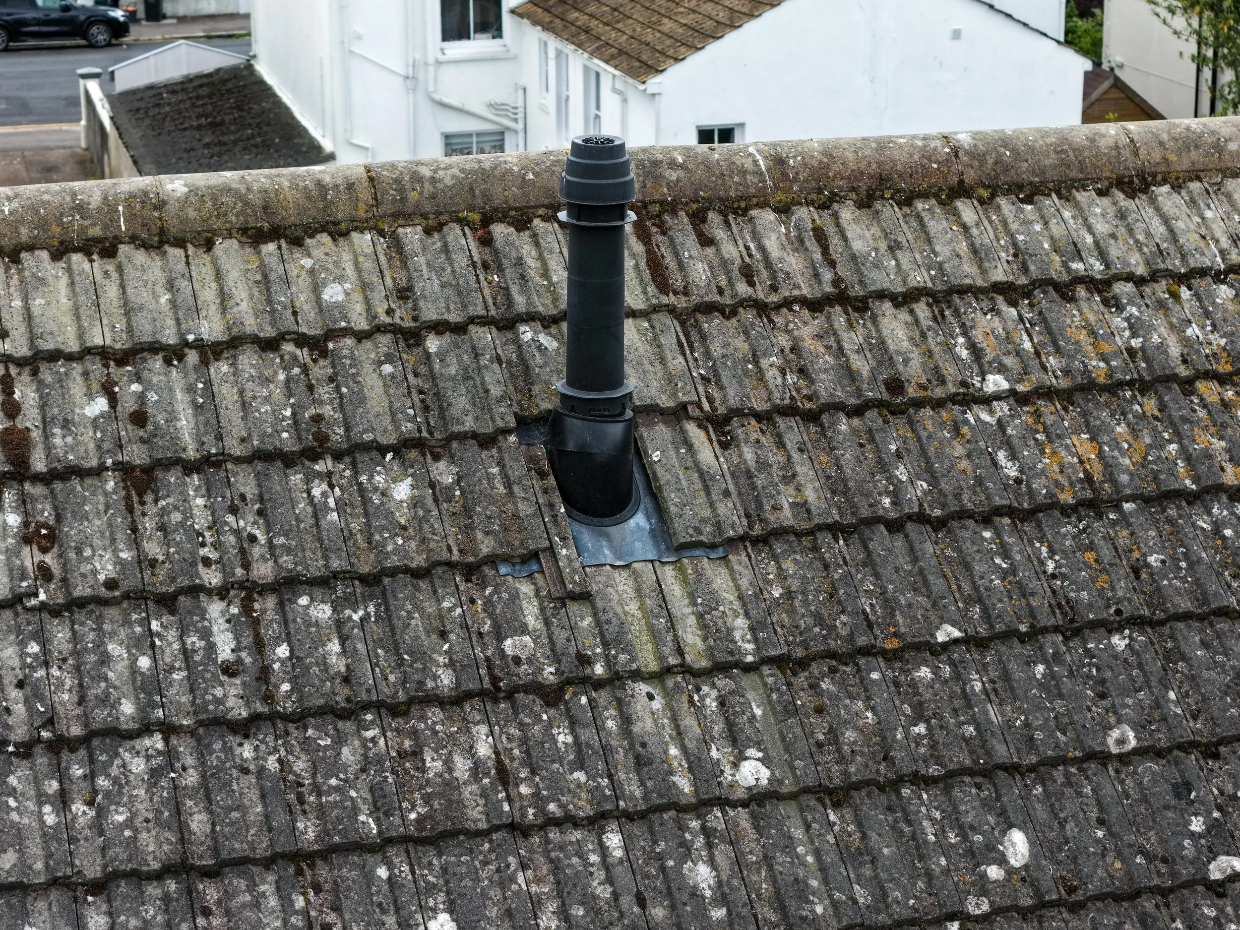 View of a rooftop with weathered, moss-covered tiles and a black vent pipe. In the background, there are white residential buildings and cars parked on the street.