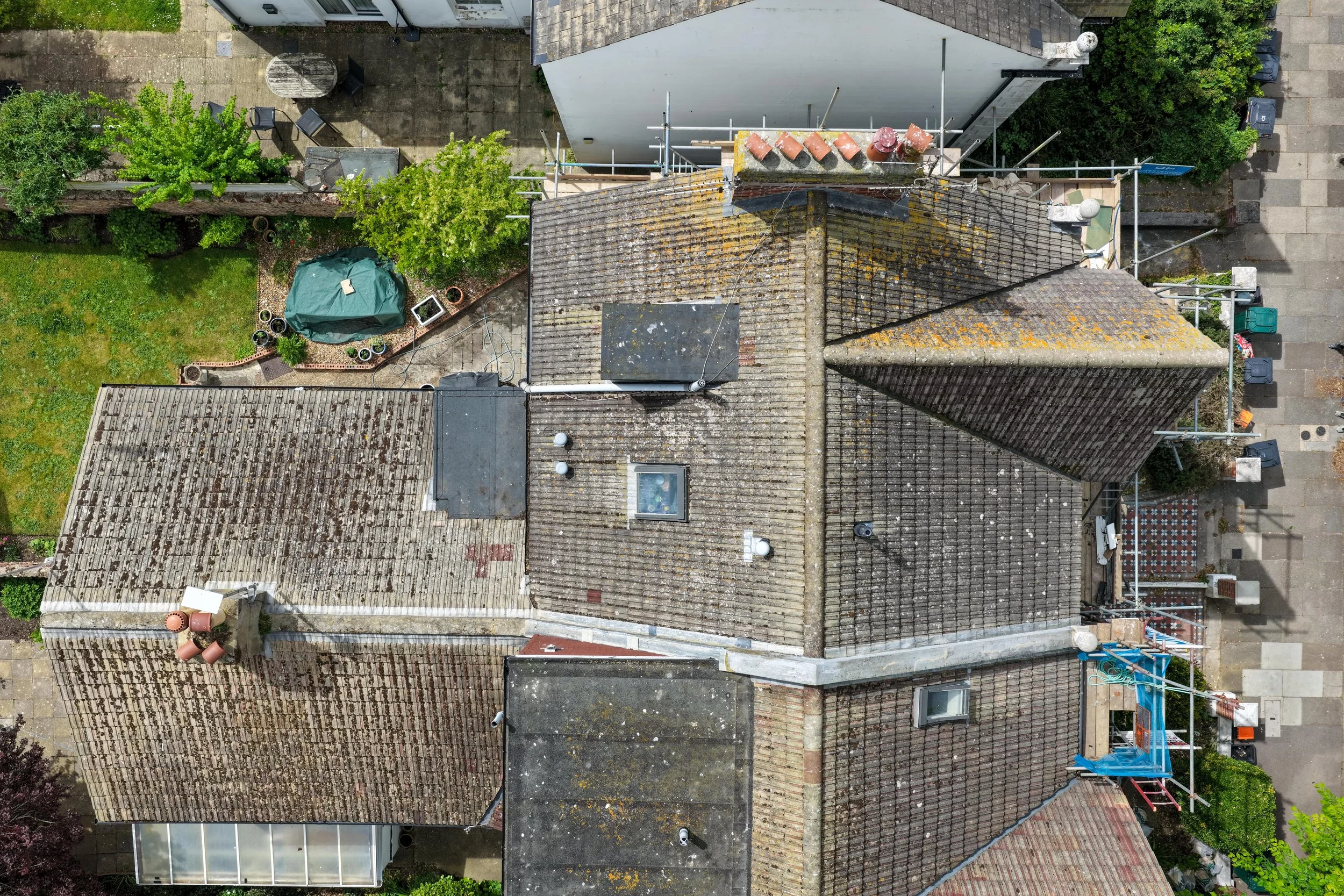 An aerial view showing rooftops, a small backyard with potted plants, green trees, patio furniture, and a paved area with trash bins and some construction scaffolding.