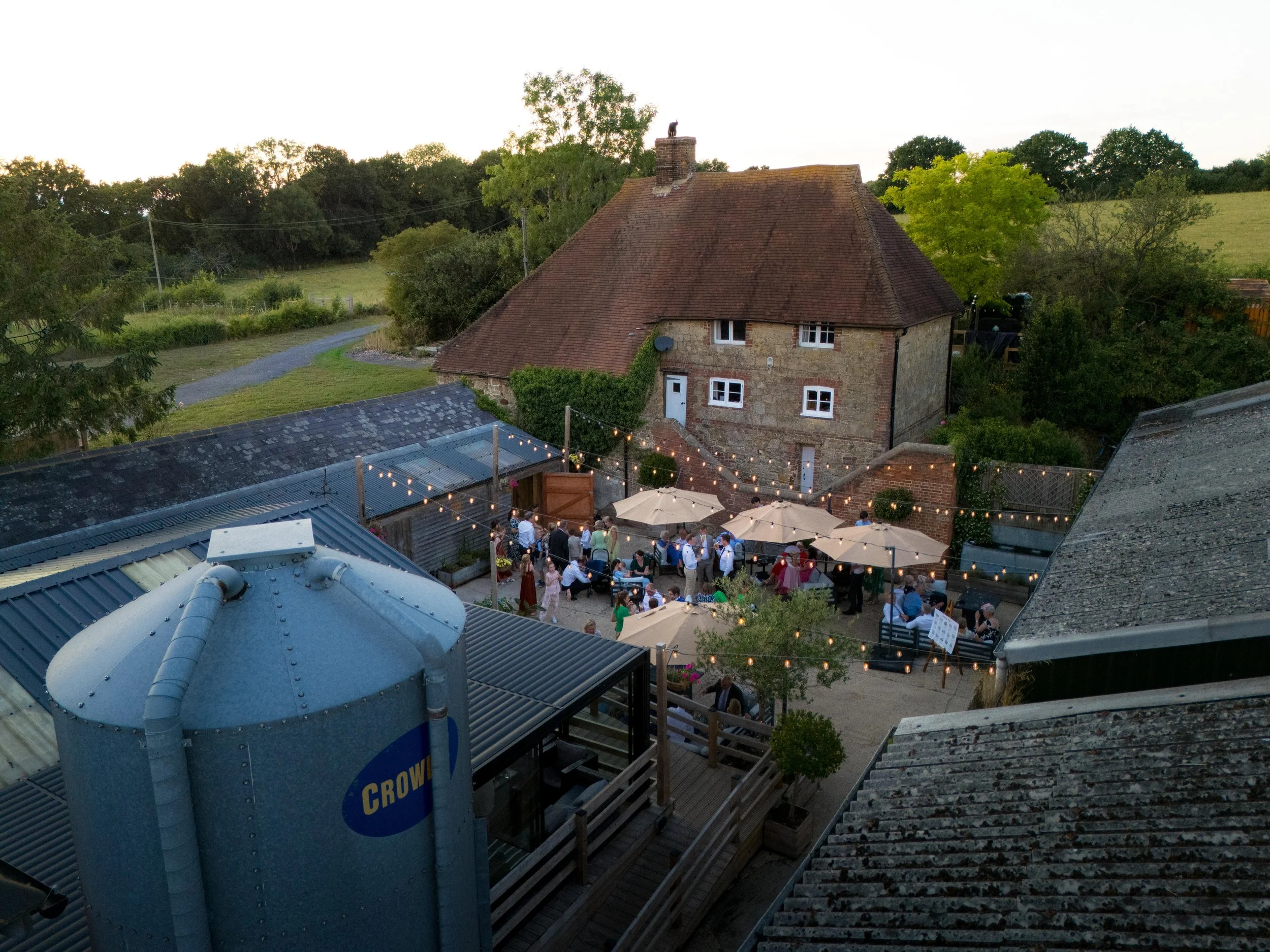 An outdoor gathering on a patio with string lights, surrounded by green trees and a rustic brick house with a sloped roof.