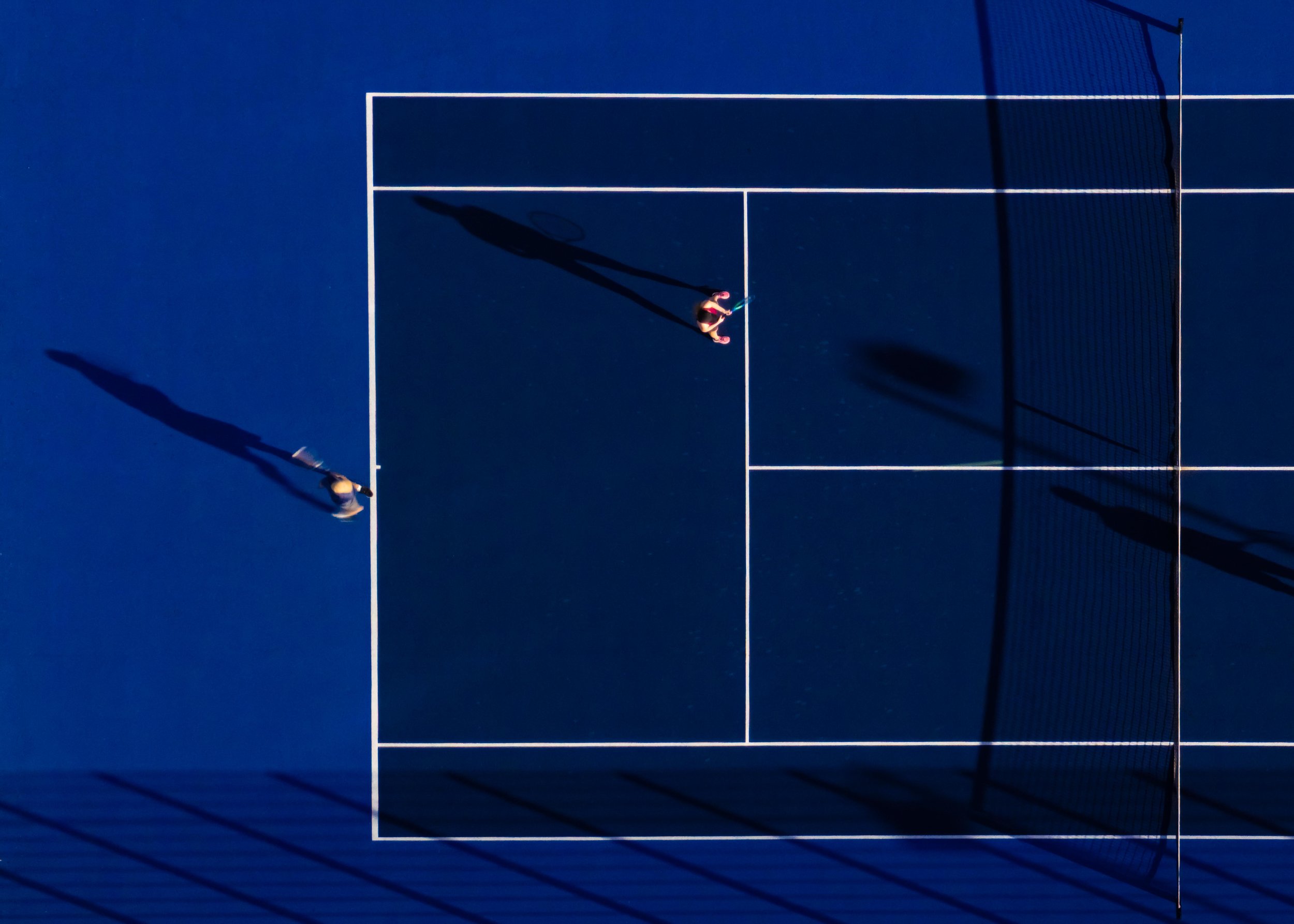 Top-down view of a tennis court with a player preparing to serve, casting a shadow, with a net on the right side.