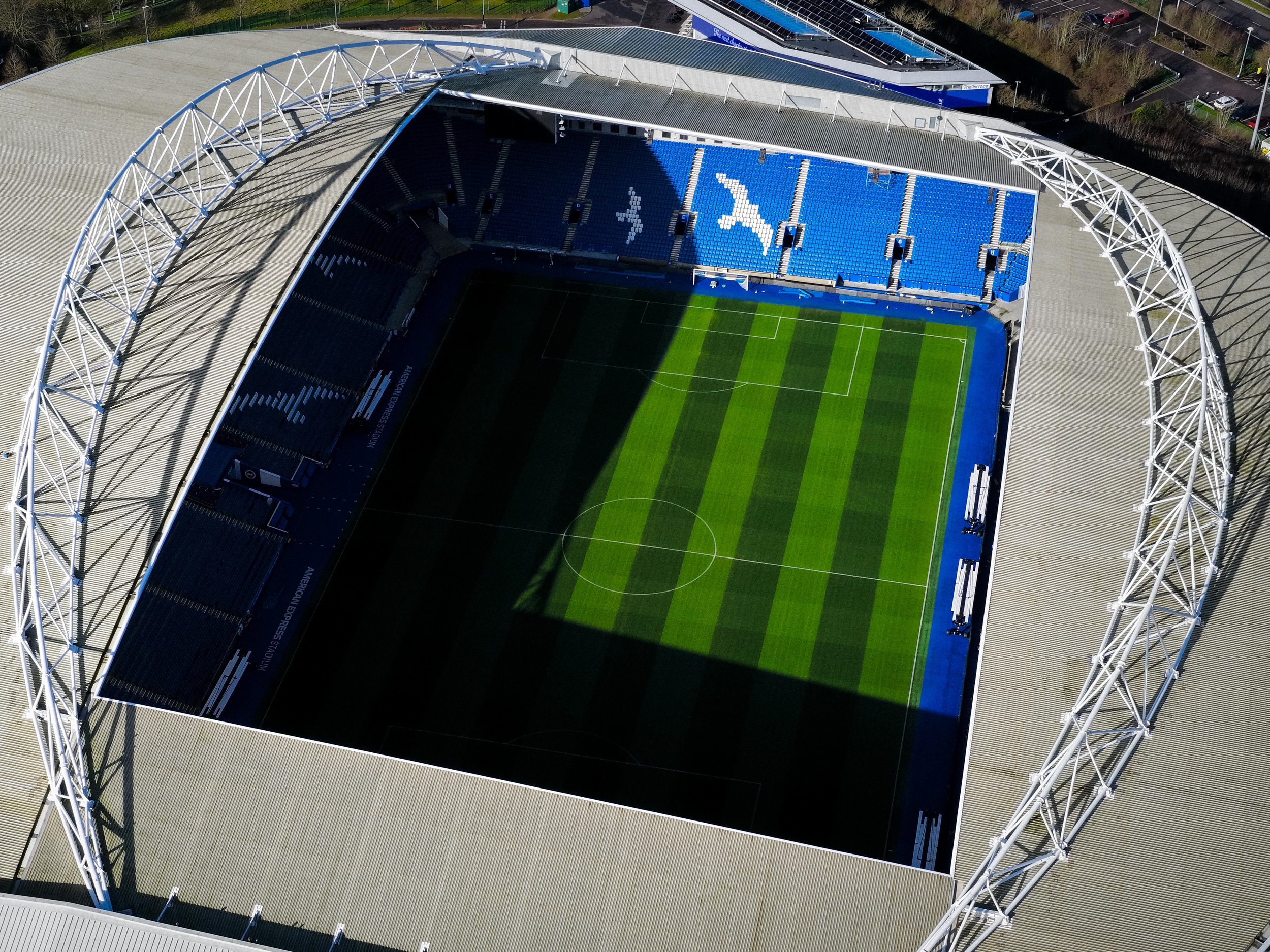  aerial view of a Brighton FC Amex football stadium with blue seating, green field, and a large white arch structure on one side.