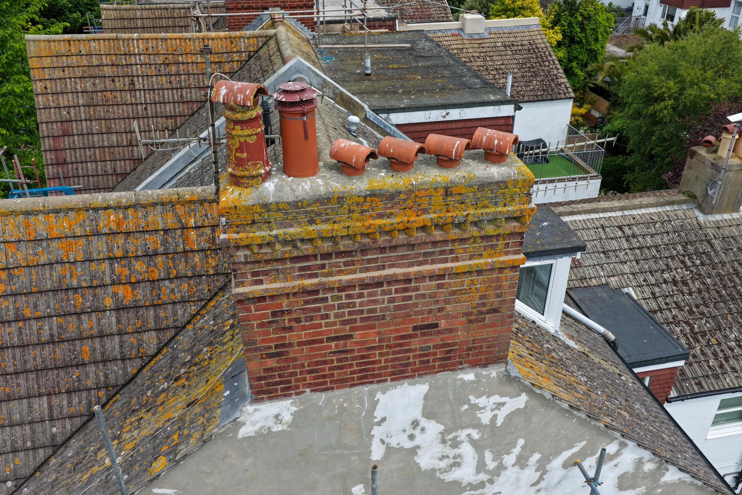 A rooftop with multiple chimneys, some covered in moss and lichen, with rusty storm caps and antenna structures. The roof has moss growth and weathering, with a neighboring white house and lush greenery in the background.