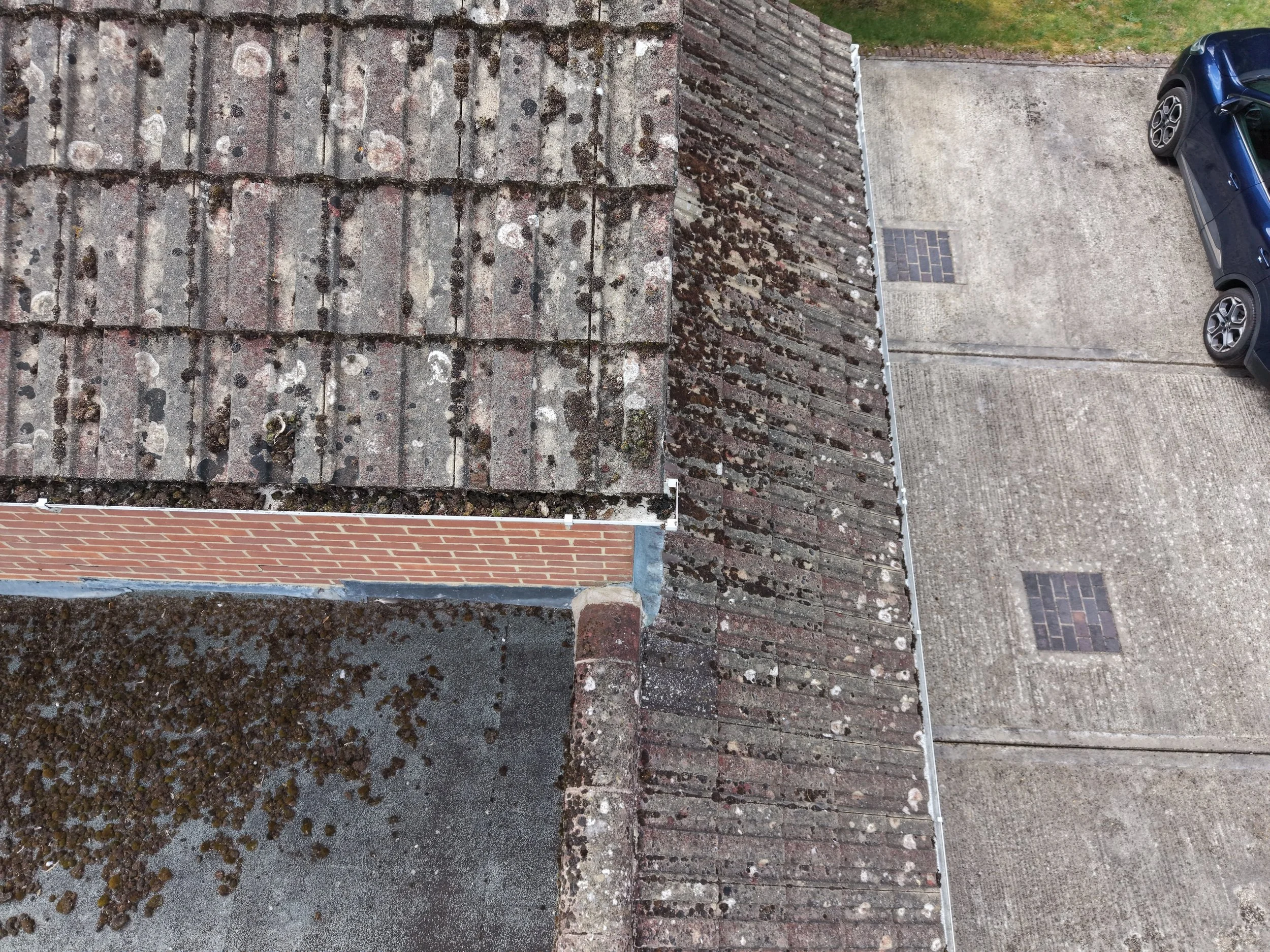 The image shows the roof of a building covered with weathered, mossy, lichen-covered shingles and a brick wall at the edge of the roof. The roof materials include both tiles and shingles, with moss growth visible on the surface. In the background, th
