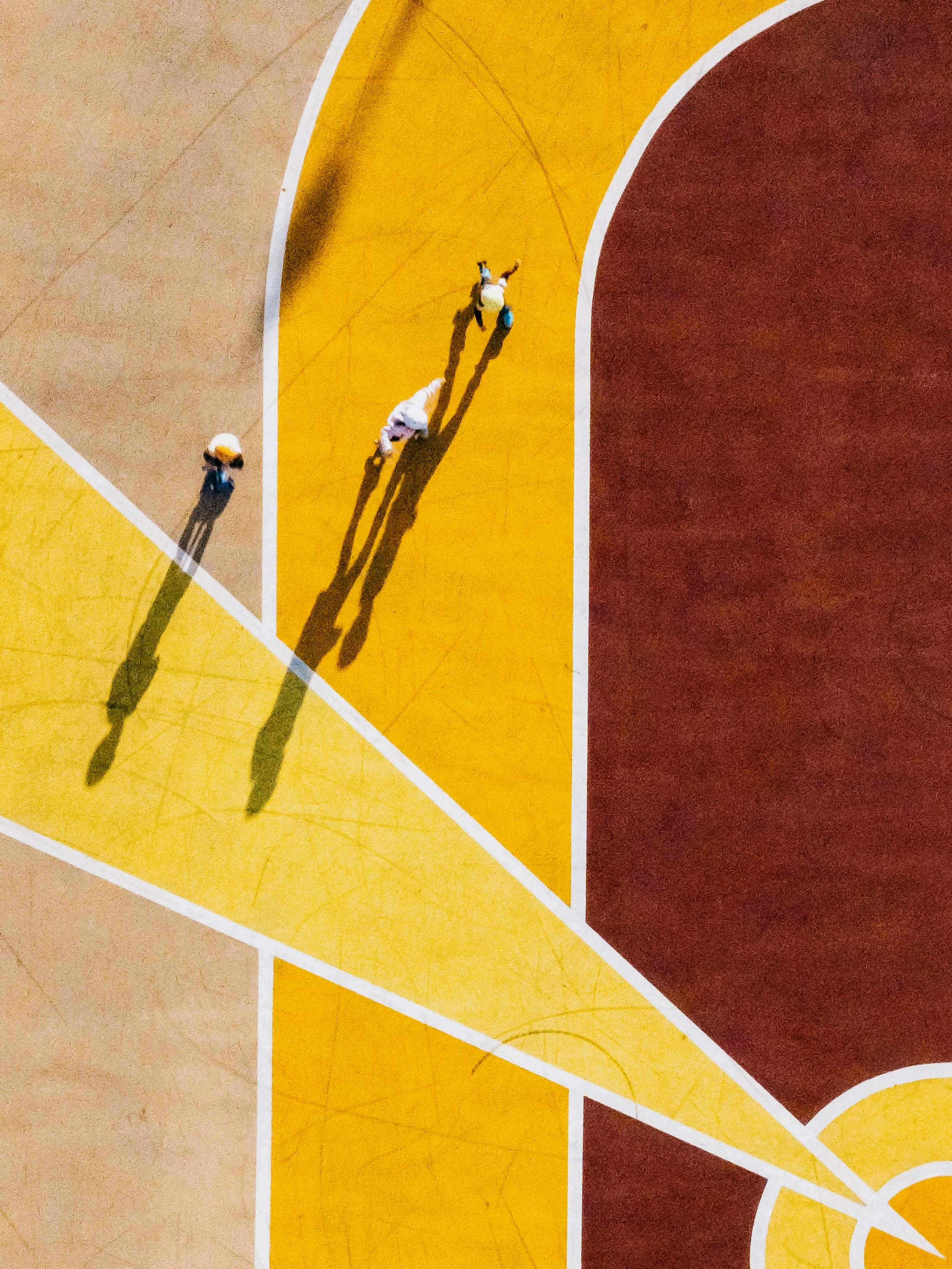 Top-down view of a skate park, showing three people skating on the yellow section, with their elongated shadows casting across the court.