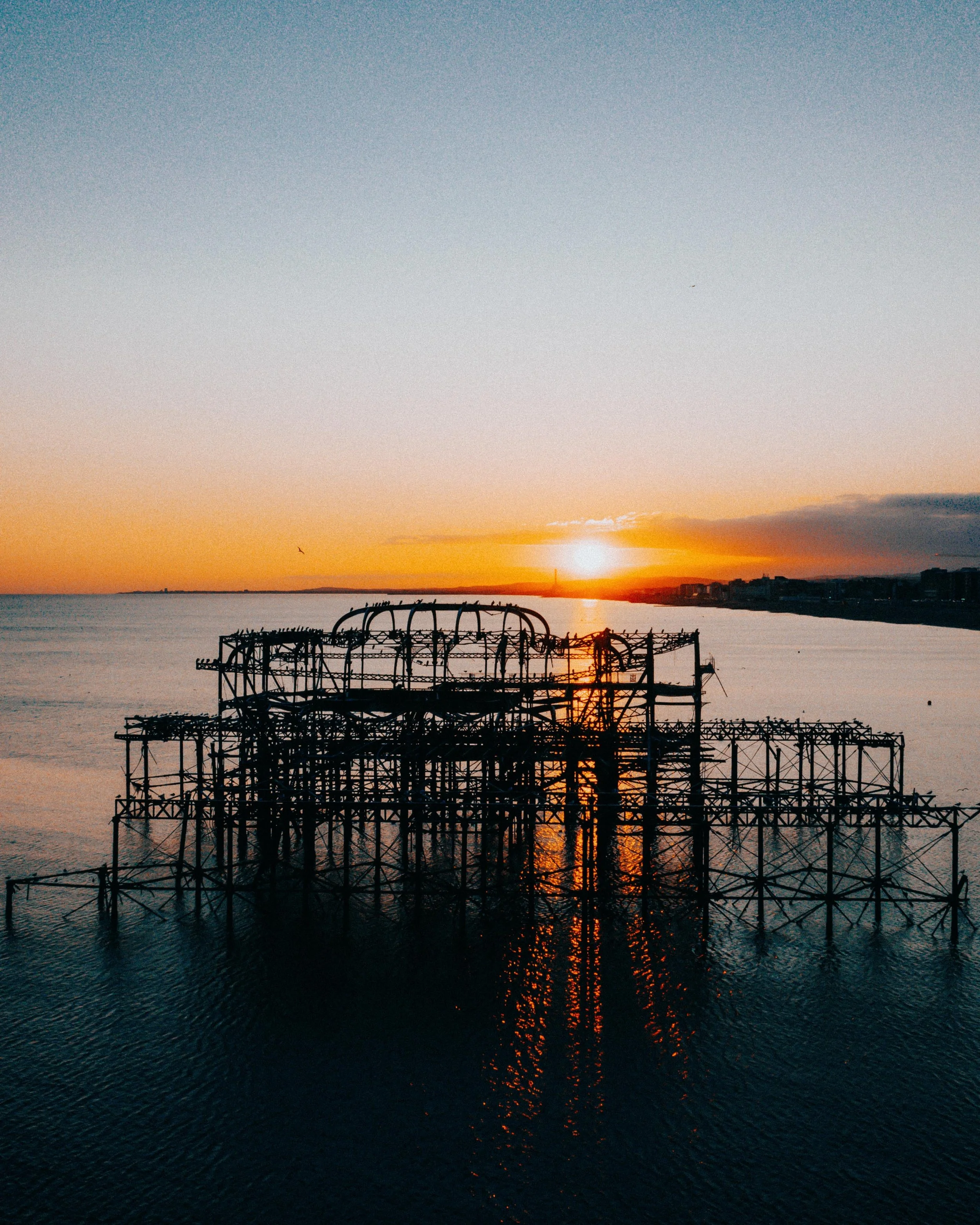 Sunset over a body of water with silhouettes of an old, dilapidated amusement park ride or structure in the foreground and a cityscape in the distance. Brighton West Pier.