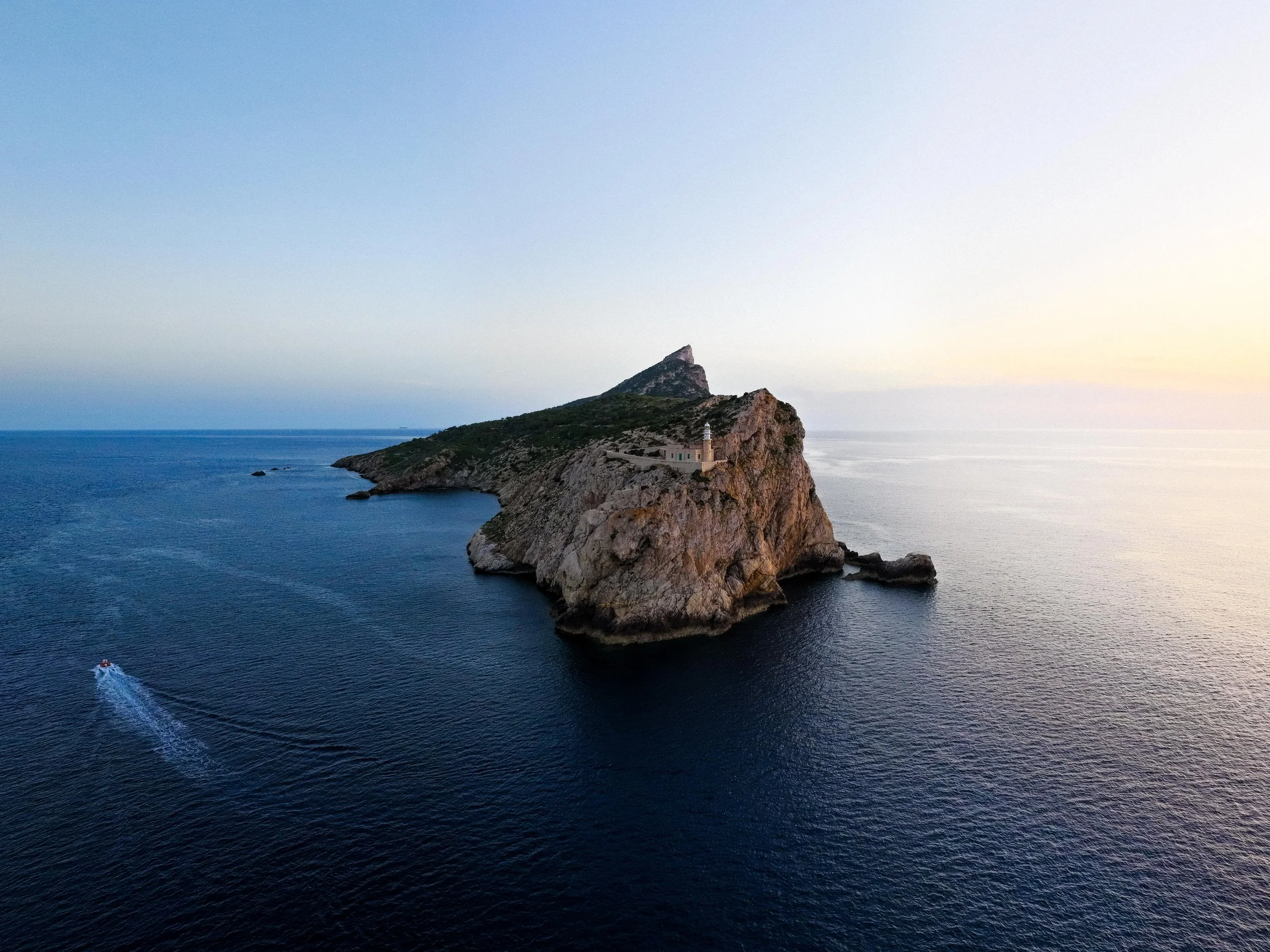 A rocky island off the coast of Majorca with a lighthouse on its edge surrounded by the ocean, with a small boat creating white wake in the water, and a mountain on the island in the background, during twilight.