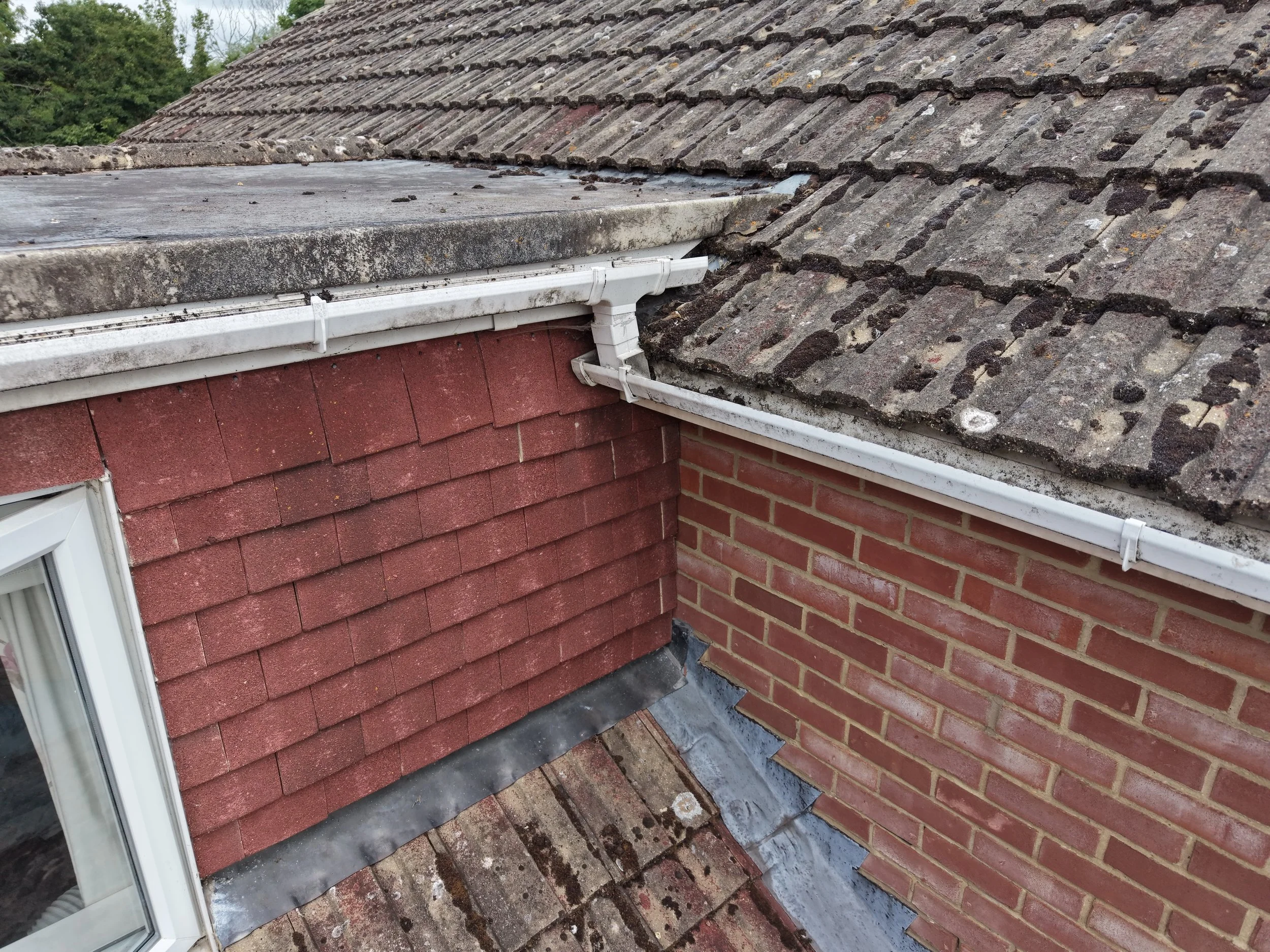 Close-up of a house's roof showing weathered gray roof tiles, a section of white gutter, and red brick walls with a window.