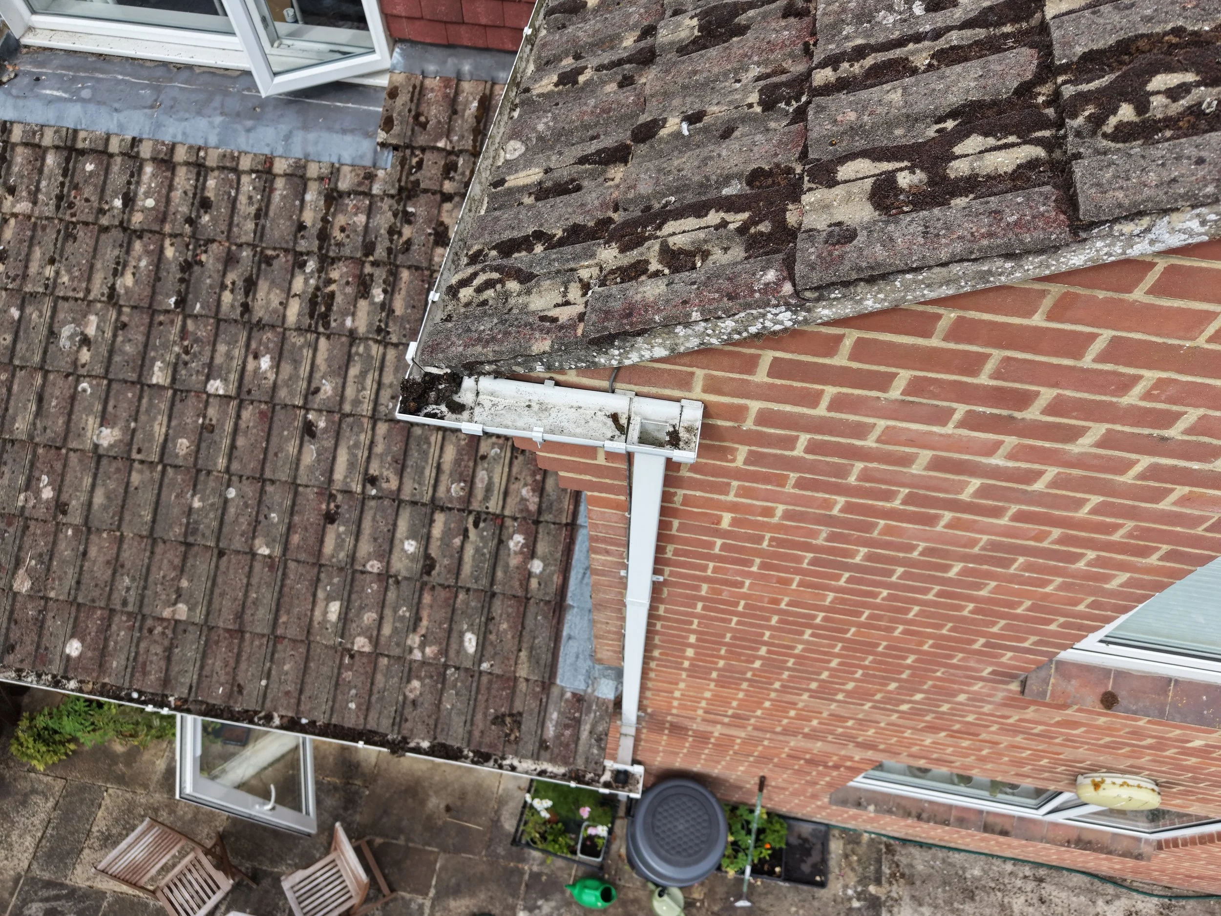 View of a rooftop with two different types of roofing materials: a traditional tile roof on the left and a flat roof with gravel on the right. There is a partially open glass door leading to a small outdoor space with flower pots, a green watering ca