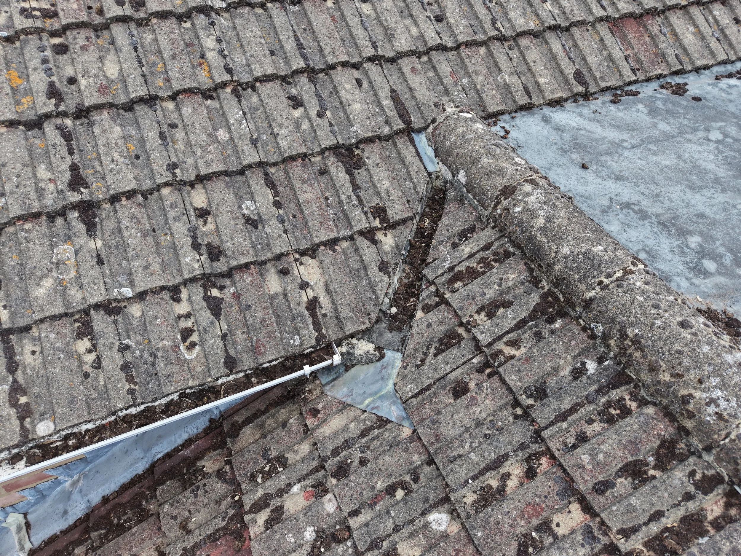 A close-up of a roof with new metal flashing installed along the edge, with weathered, mossy old tiles and a section of damaged and aged concrete or cement surface nearby.