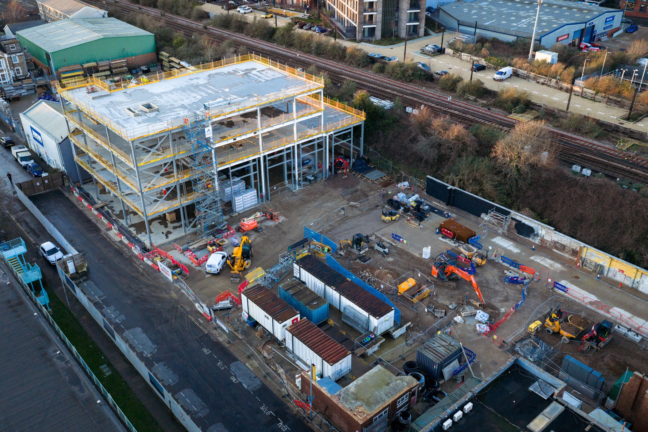 An aerial view of a construction site with a building under construction, surrounded by equipment, materials, and temporary structures. Nearby is a parking area and railway tracks, with trees and urban buildings in the background.