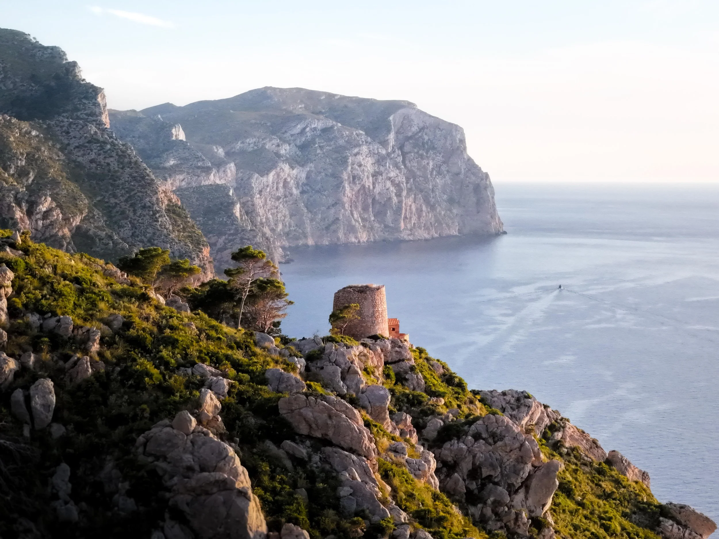 A coastal landscape in Majorca featuring steep rocky cliffs with greenery in the foreground, overlooking a calm sea with a small boat in the water.